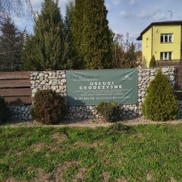 Lex Terrae Łukasz Szajewski - Baner firmy geodezyjnej Lex Terrae na ogrodzeniu z gabionów, otoczony zielenią i drzewami. Widoczny numer telefonu i adres e-mail. Słoneczny dzień.