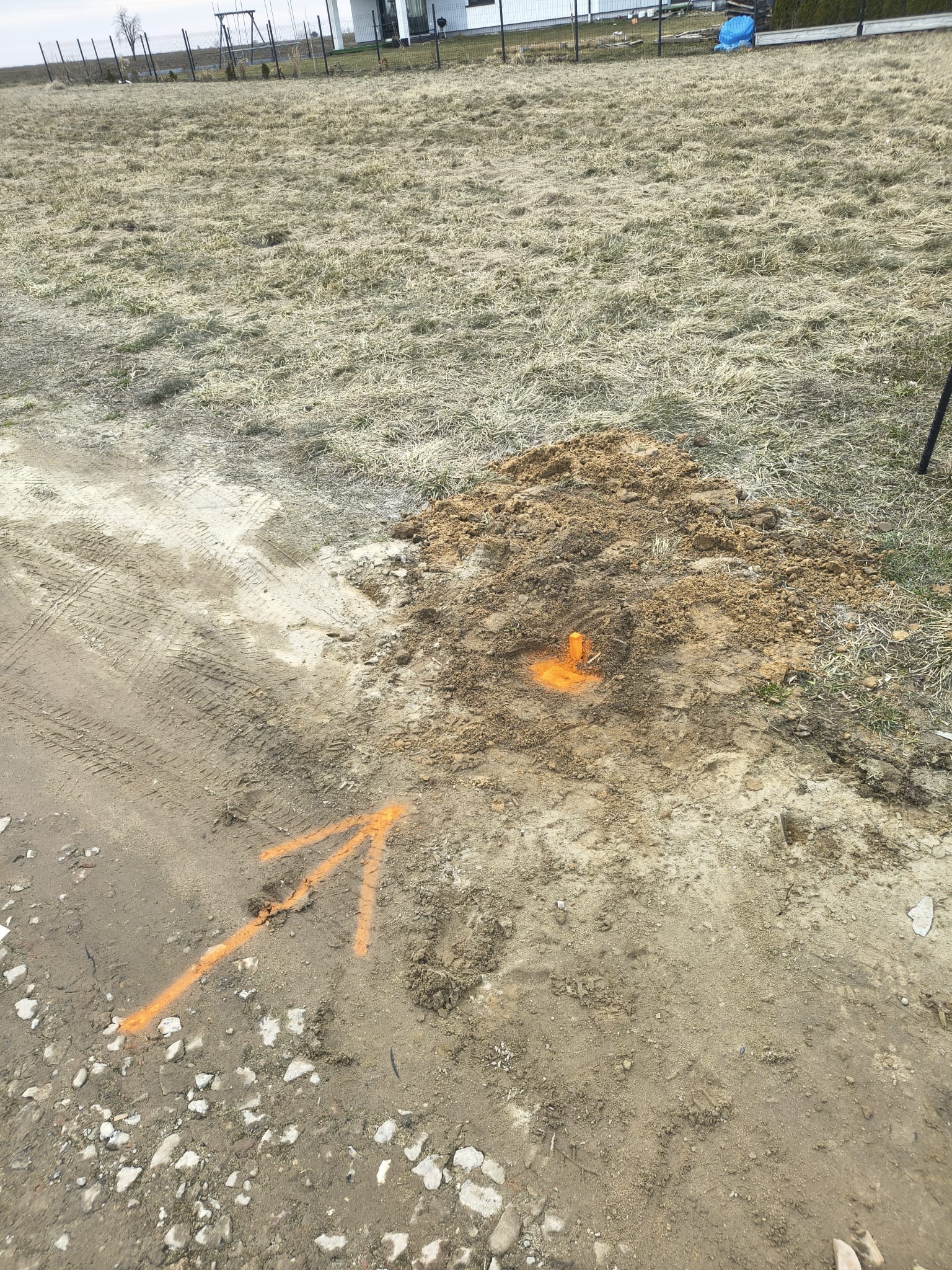 Wytyczenie działki: pomarańczowy palik w ziemi, wskazujący punkt geodezyjny. W tle widoczny dom i trawa. Ślady opon na ziemi. Pomarańczowa strzałka na ziemi.