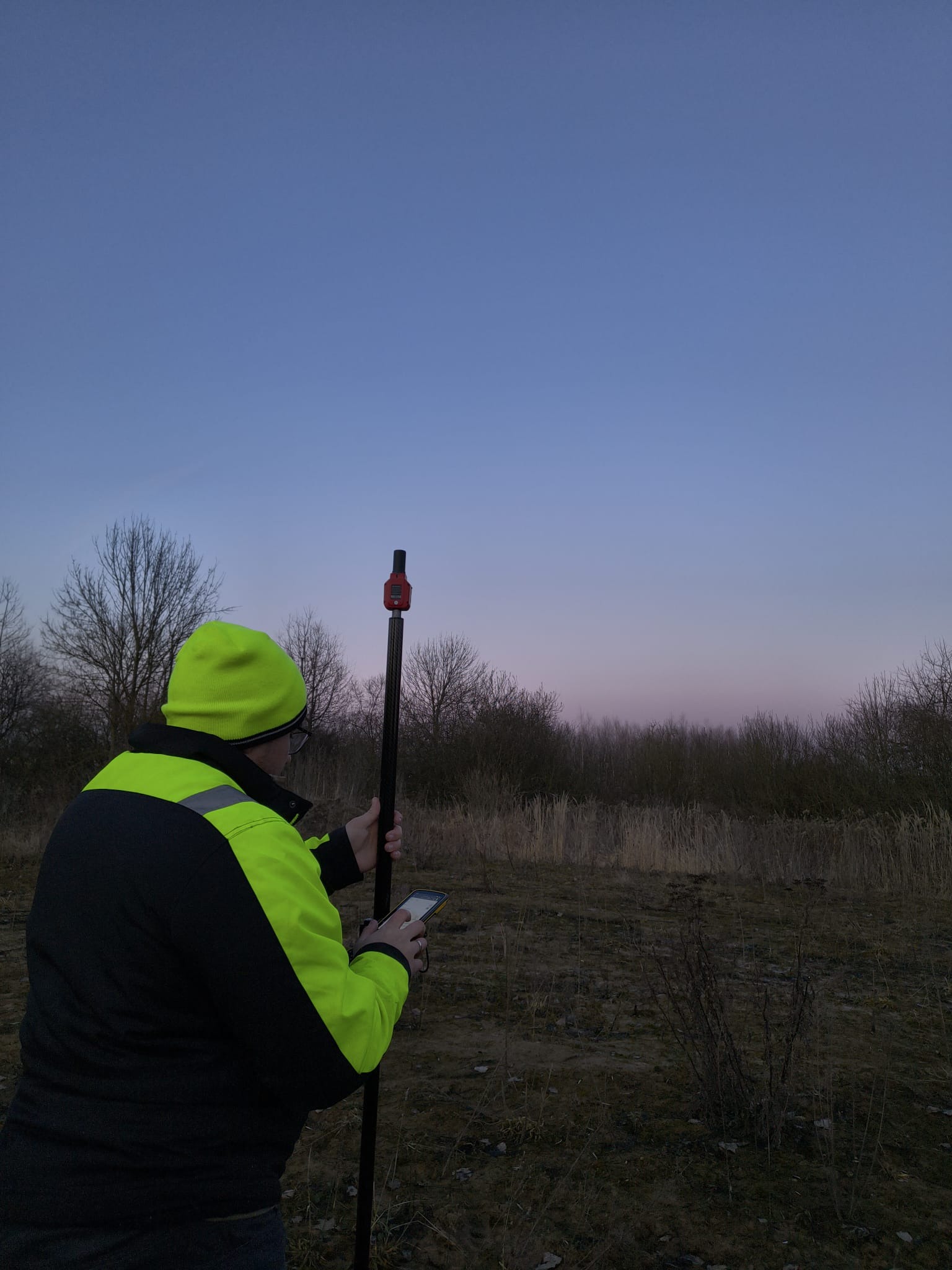 Osoba w odblaskowej kurtce i czapce, trzymająca tyczkę geodezyjną i smartfon na tle pola i drzew przy zmierzchu. Pomiar terenu w trakcie prac.