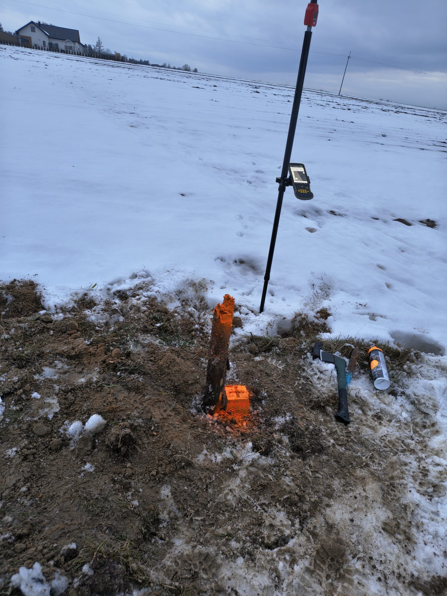 Zimowy krajobraz z wytyczonym punktem pomiarowym, oznaczonym palikiem i pomalowanym na pomarańczowo, obok leży toporek i farba w sprayu. W tle pole i dom.