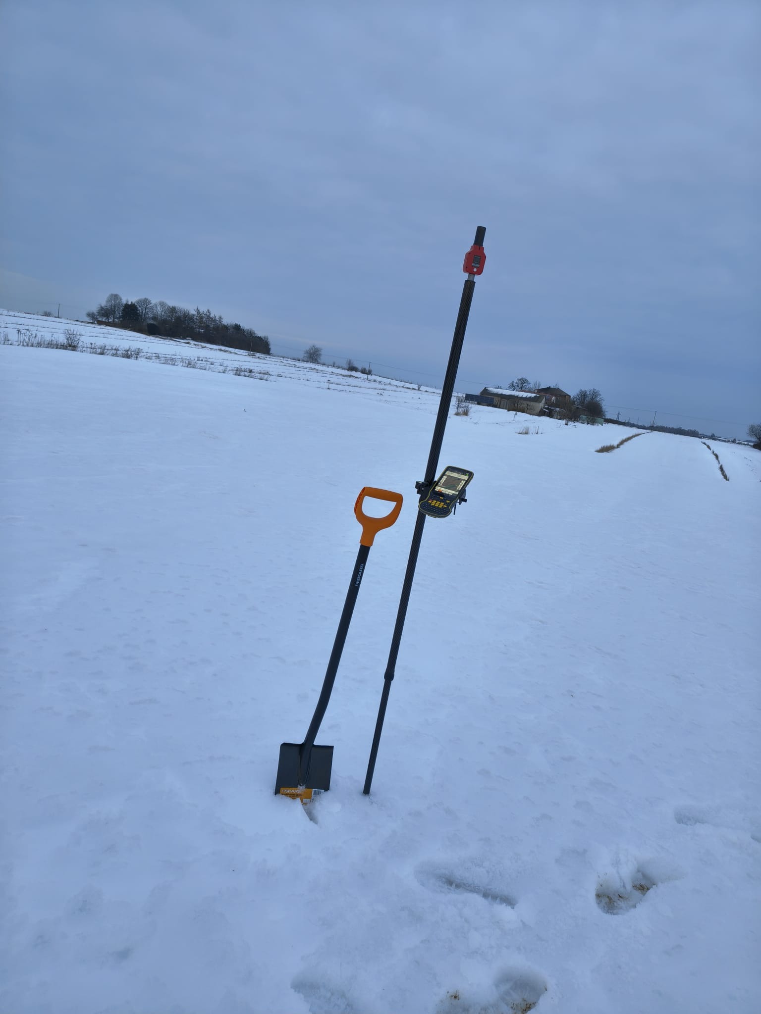 Zimowy krajobraz z łopatą Fiskars i tyczką geodezyjną z odbiornikiem GPS na zasypanym śniegiem polu. W tle widoczne zabudowania.