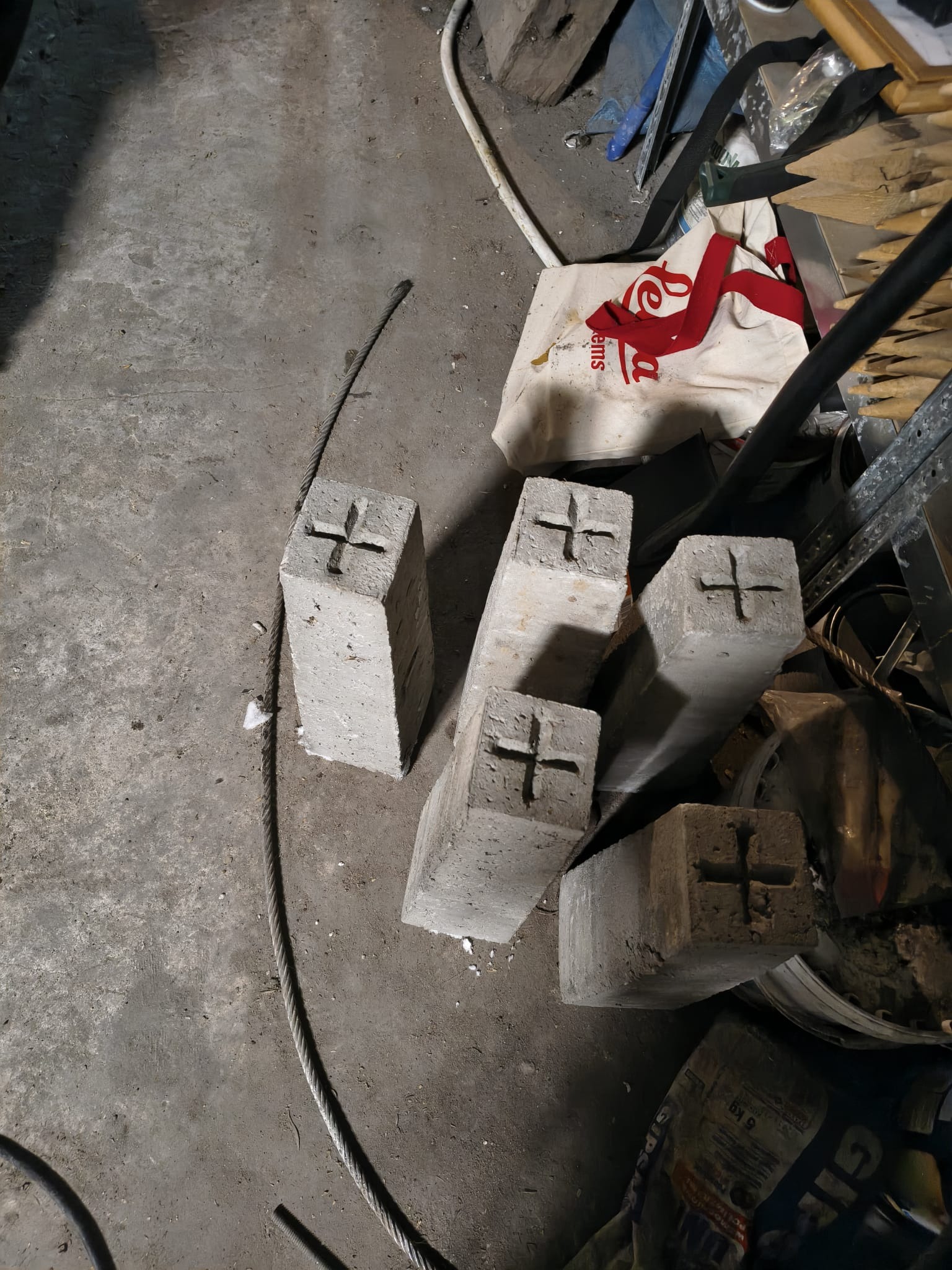Cztery betonowe słupki geodezyjne z wyżłobionym krzyżem na szczycie, leżące na betonowej podłodze w otoczeniu narzędzi i materiałów budowlanych.