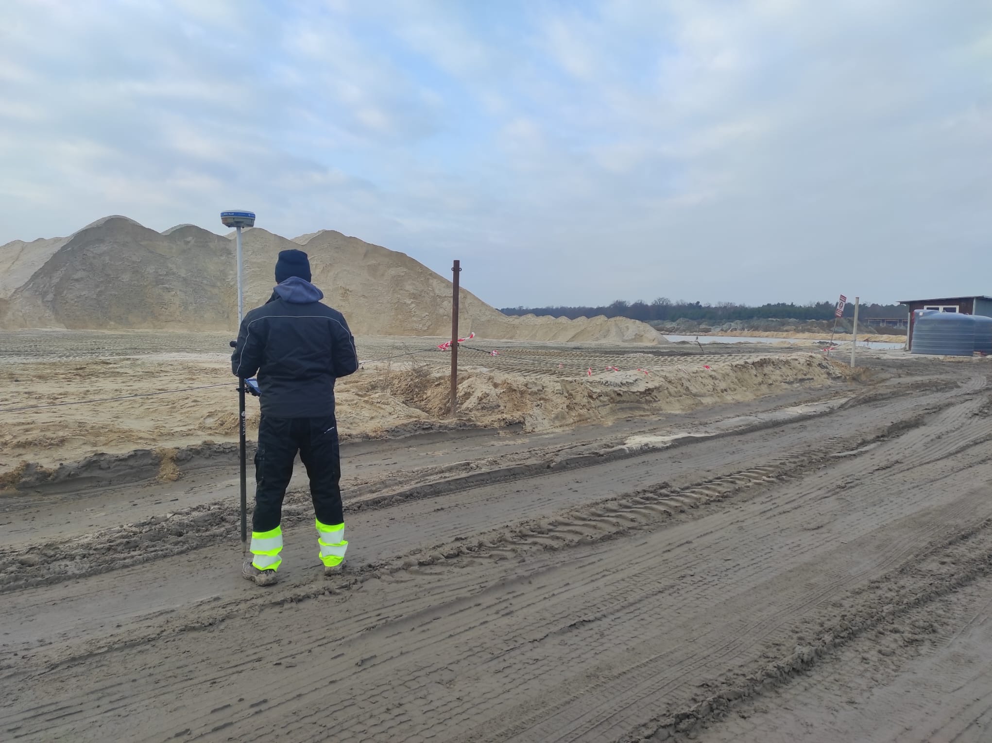 Praca pomiarowa w Świdniku Dużym Drugim: osoba z tyłu, w odzieży roboczej, używa sprzętu geodezyjnego na tle hałd piasku i nieba.
