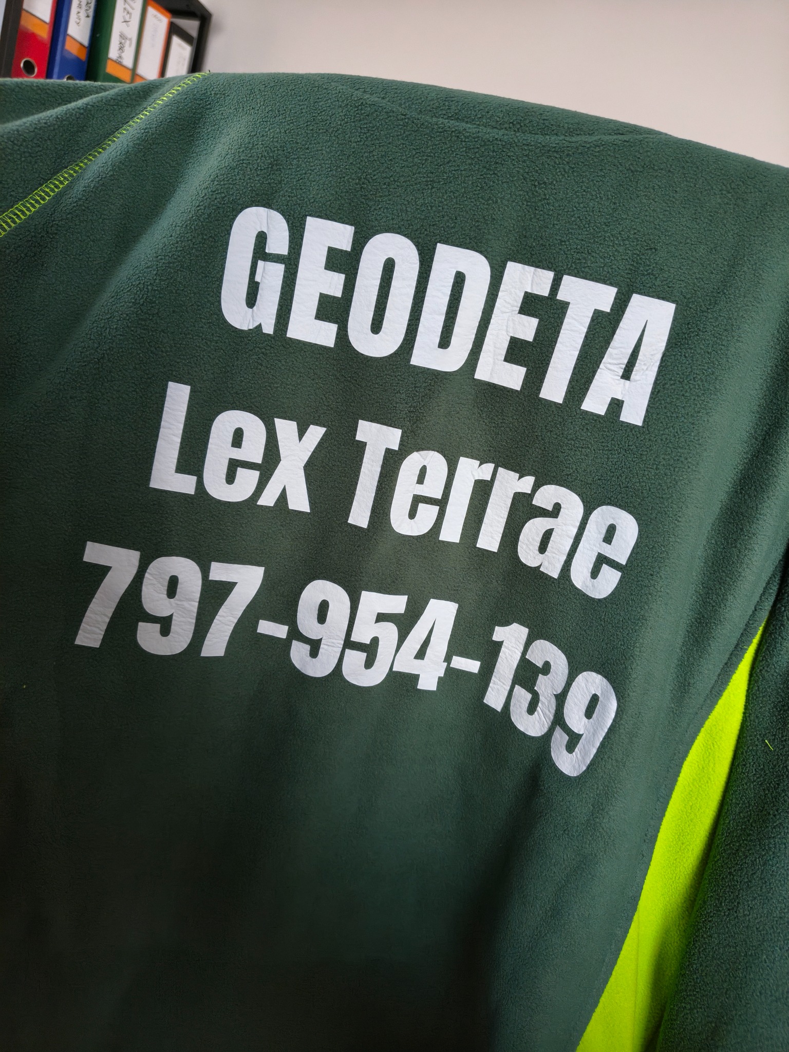 Zielona bluza polarowa z nadrukowanym białym napisem 'Geodeta Lex Terrae 797-954-139', widoczna część rękawa w kolorze jaskrawej zieleni.