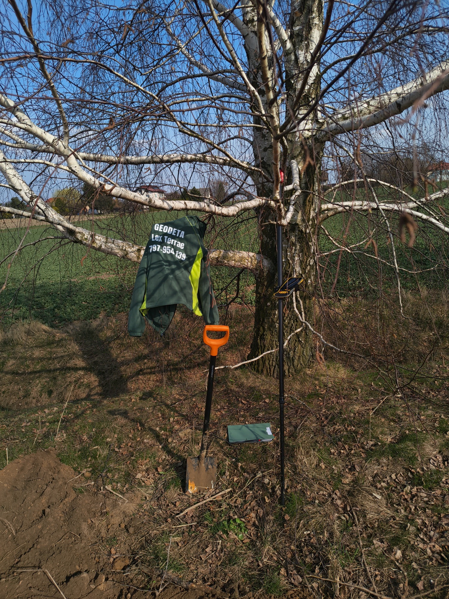 Sprzęt pomiarowy i odzież robocza firmy geodezyjnej zawieszone na brzozie na tle pola. Widoczna łopata i notatnik. Słoneczny dzień.
