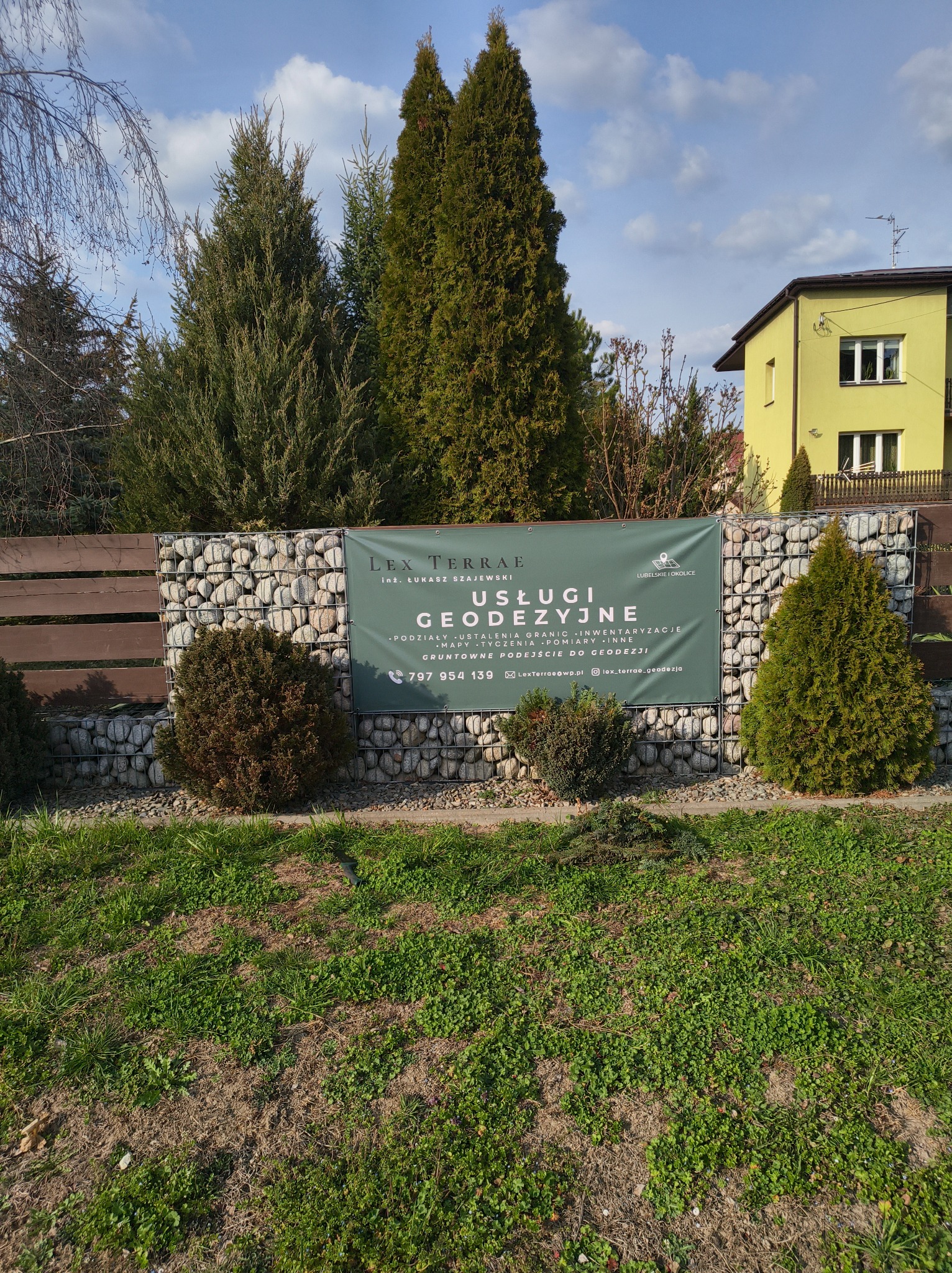 Baner firmy geodezyjnej Lex Terrae na ogrodzeniu z gabionów, otoczony zielenią i drzewami. Widoczny numer telefonu i adres e-mail. Słoneczny dzień.