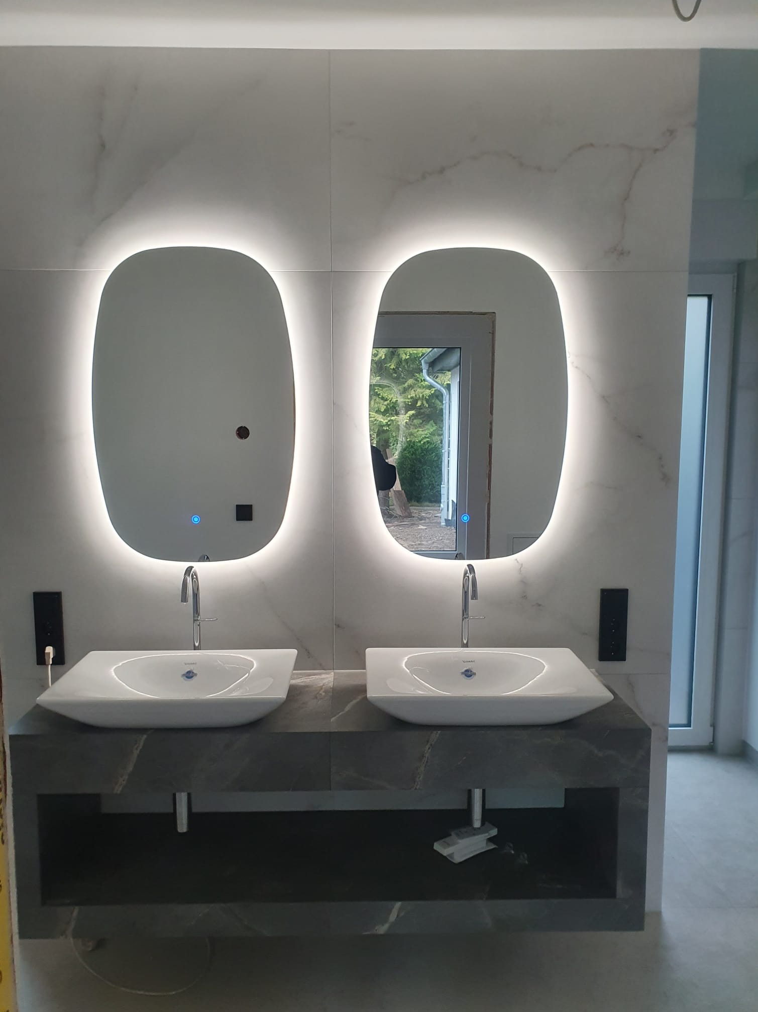 Nowoczesna łazienka z dwoma umywalkami nablatowymi i podświetlanymi lustrami o zaokrąglonym kształcie. Marmurowe płytki na ścianach i szara szafka pod umywalkami.