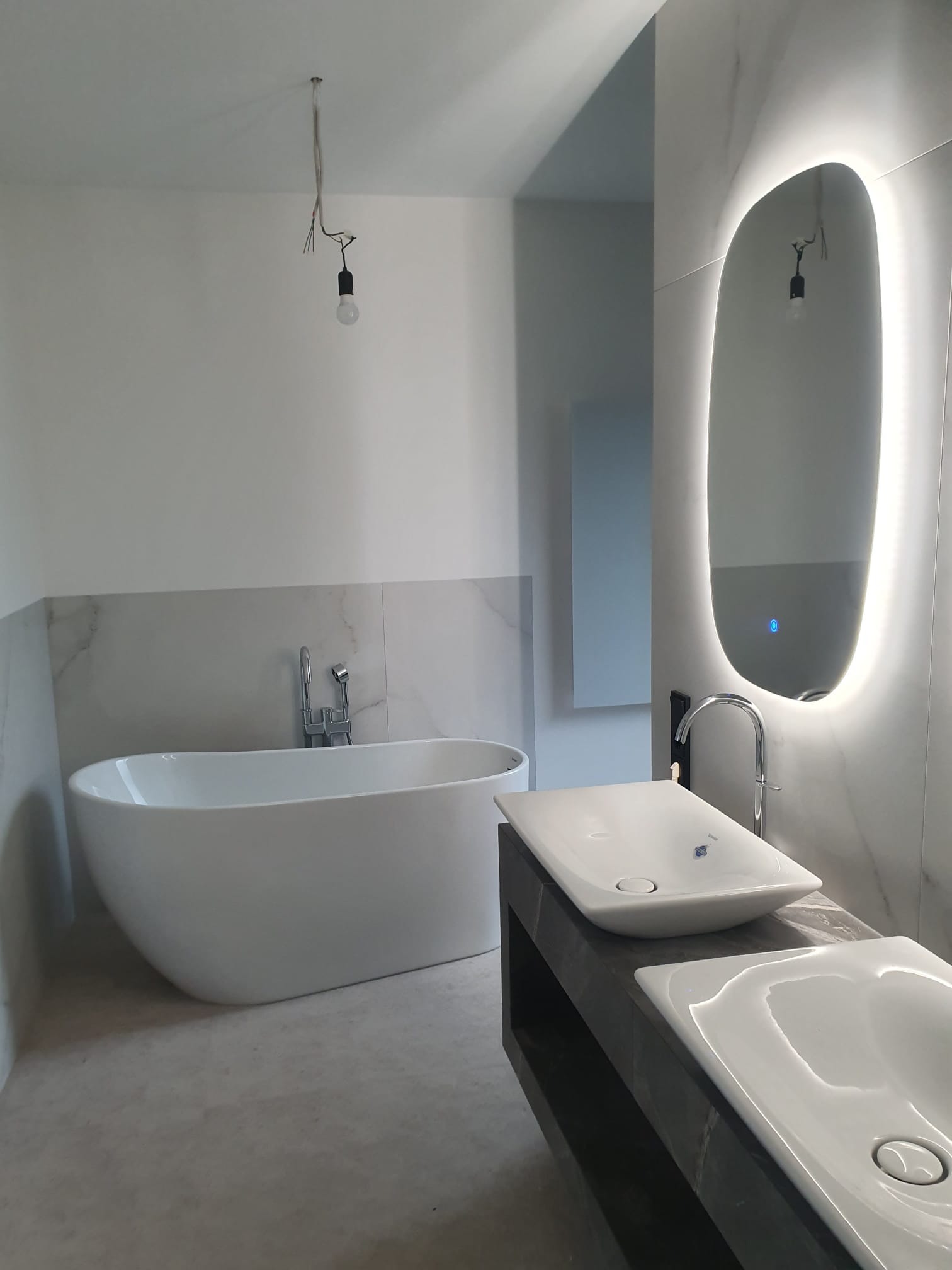 Nowoczesna łazienka w trakcie wykańczania: wolnostojąca wanna, podwójna umywalka nablatowa, podświetlane lustro. Widoczne niedokończone oświetlenie sufitowe.