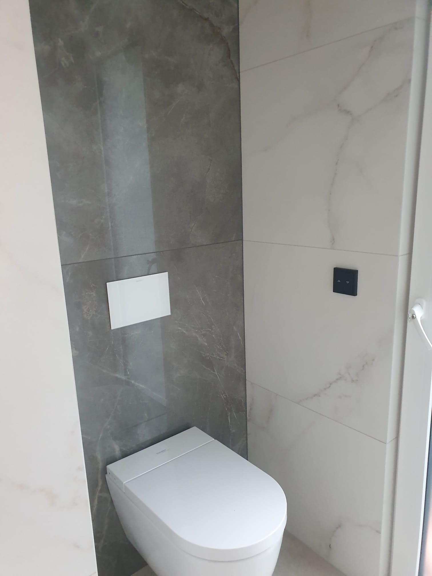 Nowoczesna toaleta z białymi i szarymi płytkami imitującymi marmur. Widoczny przycisk spłuczki i panel sterowania. Minimalistyczny design.