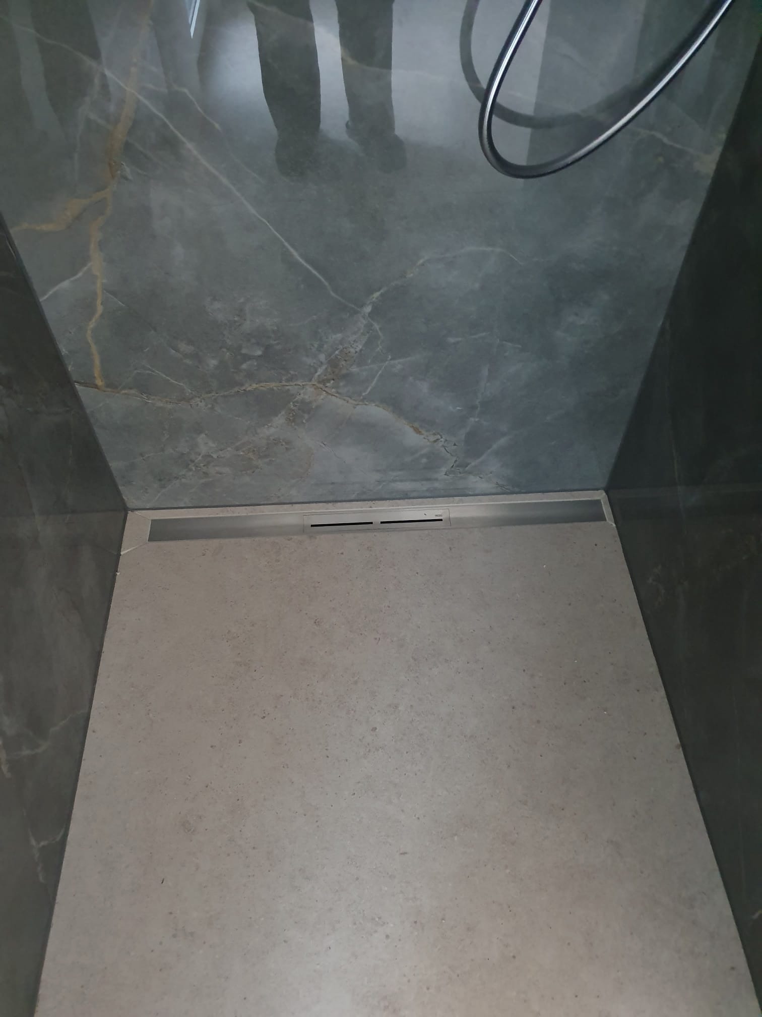 Nowoczesny odpływ liniowy w kabinie prysznicowej z szarymi płytkami imitującymi marmur z delikatnym złotym żyłkowaniem na ścianach i podłodze.