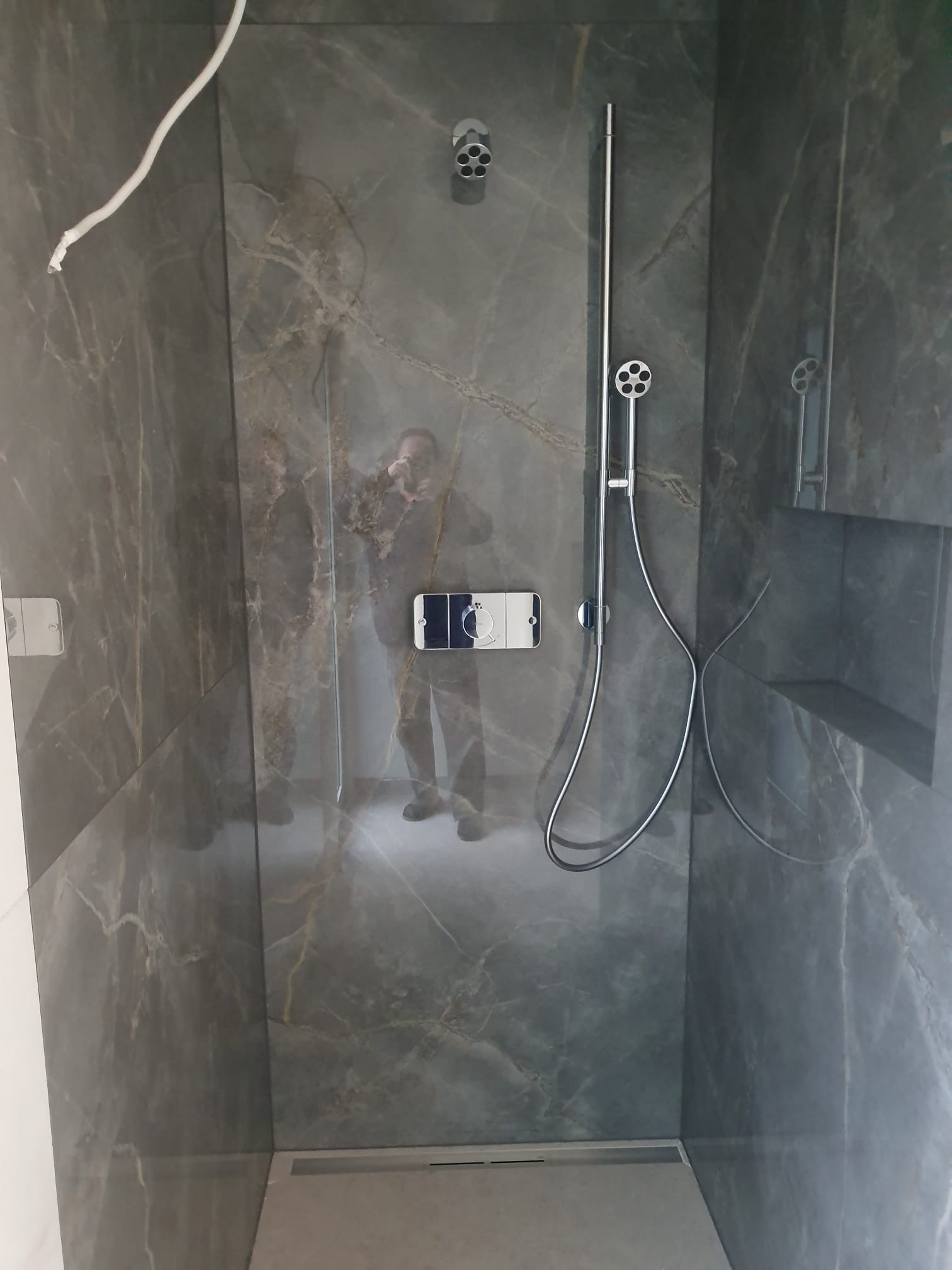 Nowoczesna kabina prysznicowa z szarymi płytkami imitującymi marmur, chromowaną armaturą i odpływem liniowym. Widoczne odbicie osoby robiącej zdjęcie.