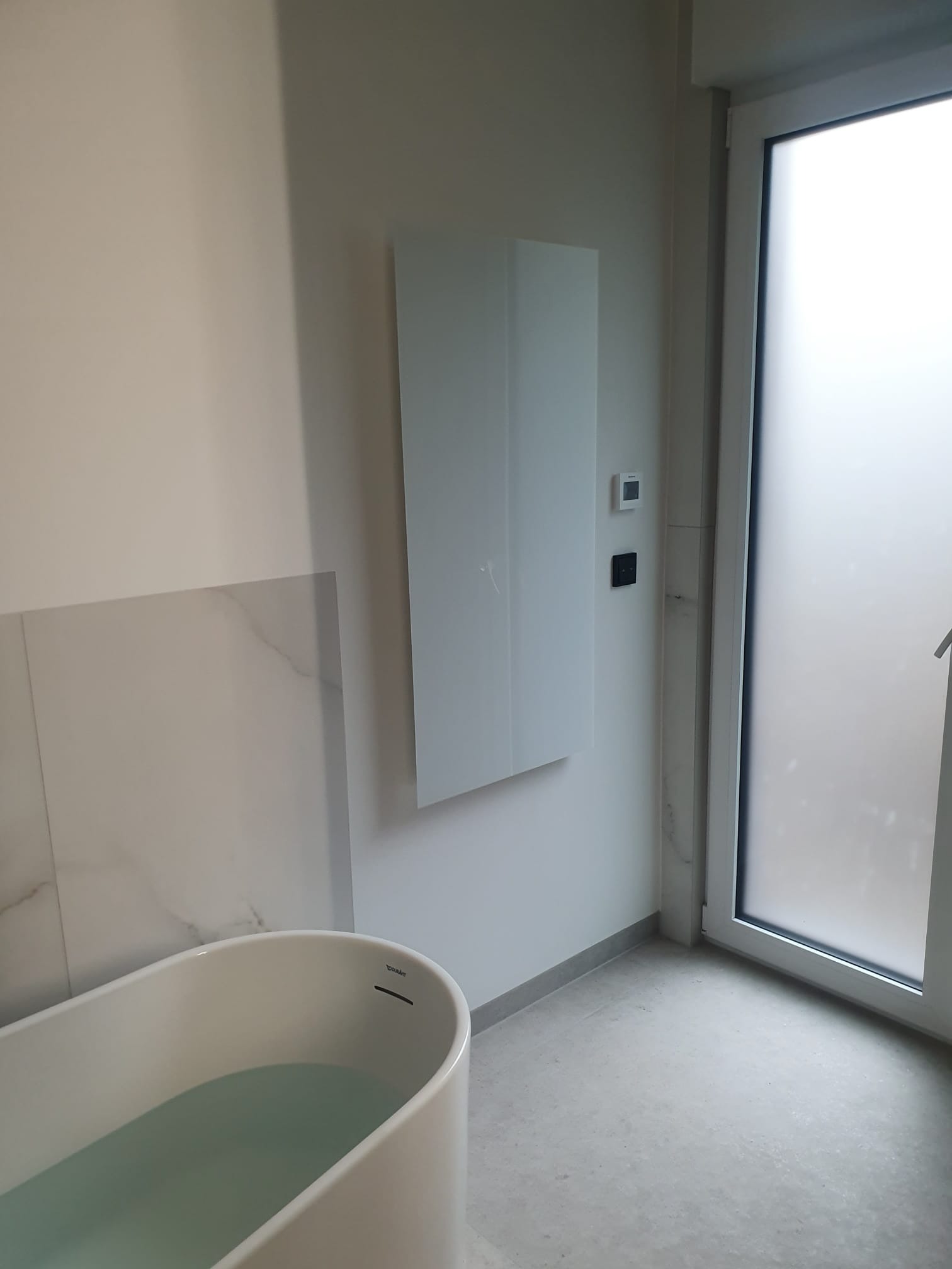 Minimalistyczna łazienka z wolnostojącą wanną Duravit, szklanym panelem grzewczym na ścianie i matową szybą okienną. Jasne płytki i neutralna kolorystyka.