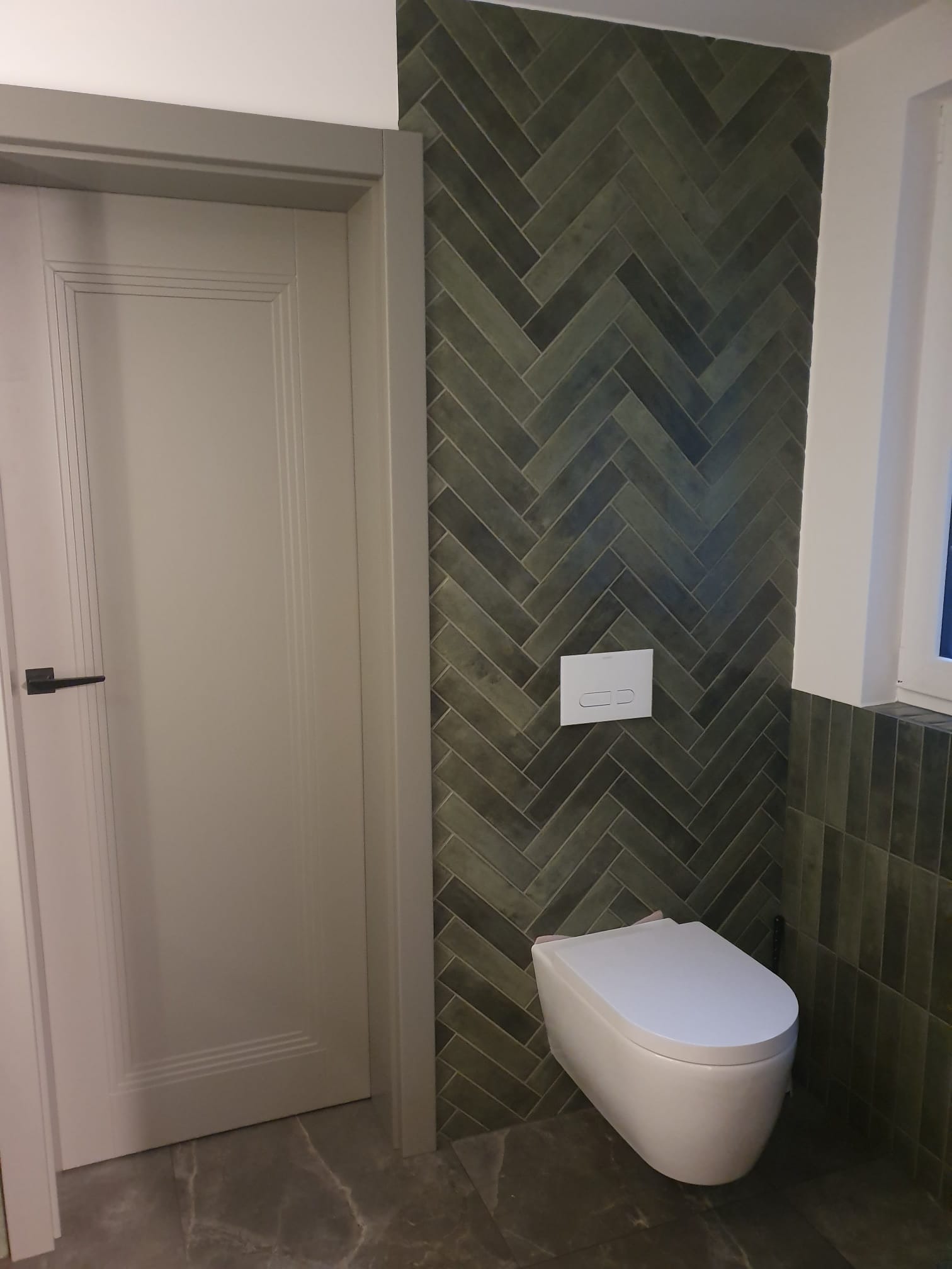 Nowoczesna toaleta podwieszana na tle ściany z zielonej, ułożonej w jodełkę glazury. Obok widoczna szara ościeżnica i białe drzwi z czarną klamką.