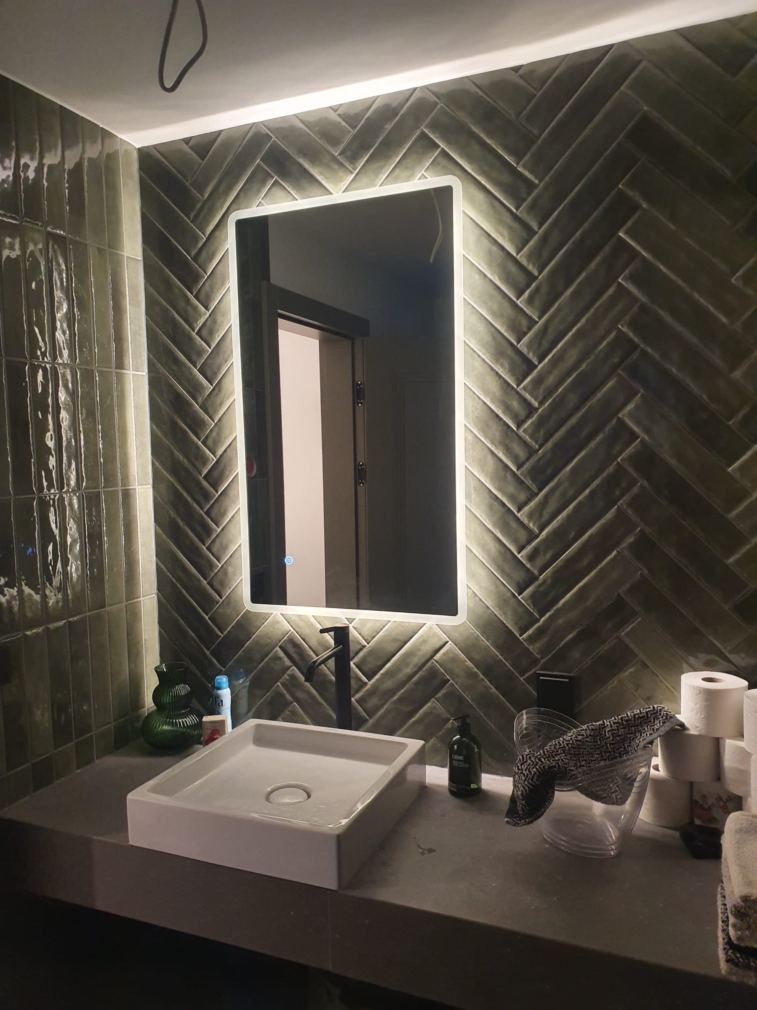 Nowoczesna łazienka z zielonymi, błyszczącymi płytkami ułożonymi w jodełkę. Umywalka nablatowa, podświetlane lustro i czarna bateria tworzą elegancki wystrój.