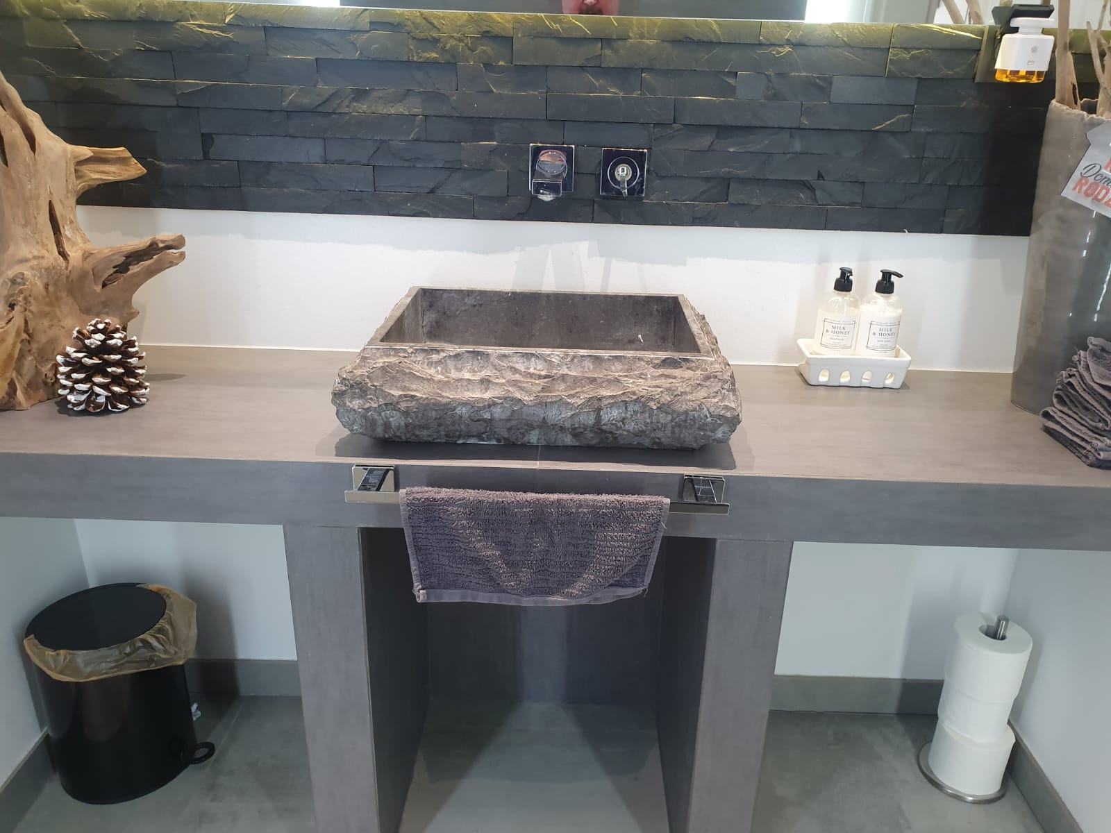 Nowoczesna łazienka z kamienną umywalką i czarną mozaiką na ścianie. Elementy dekoracyjne: drewno, szyszka, dozowniki mydła. Minimalistyczny design.