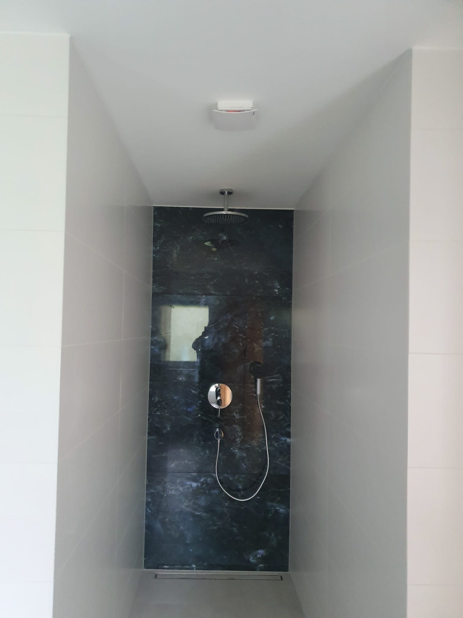 Nowoczesna kabina prysznicowa z czarną, marmurkową ścianą i deszczownicą. Jasne, kwadratowe płytki na pozostałych ścianach. Minimalistyczny design.