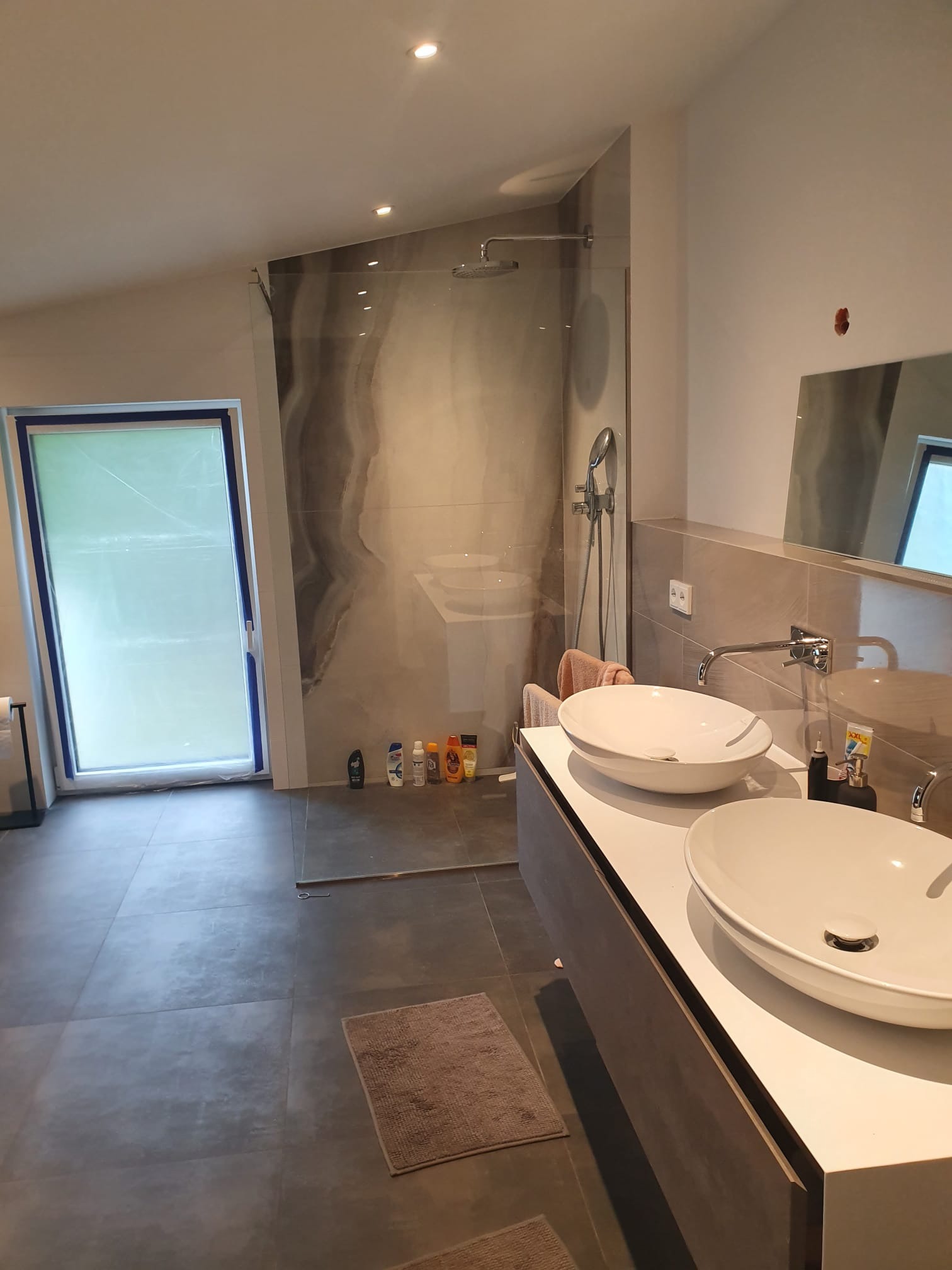 Nowoczesna łazienka na poddaszu z kabiną typu walk-in, dwiema umywalkami nablatowymi i dużymi szarymi płytkami podłogowymi. Okno zabezpieczone folią.