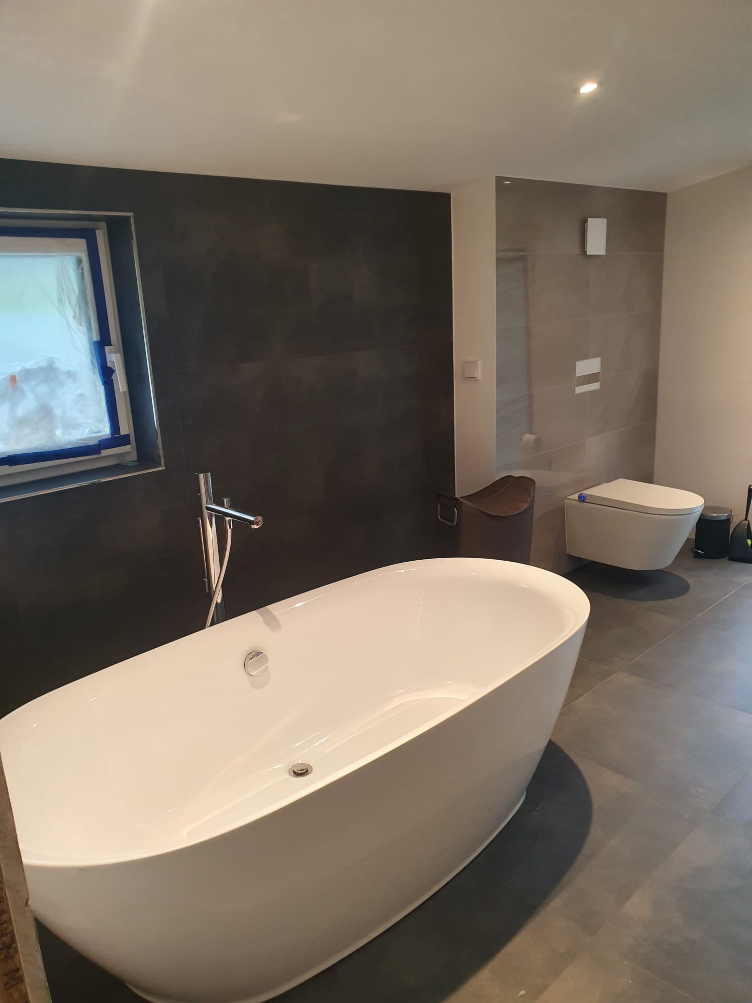 Nowoczesna łazienka z wolnostojącą wanną na tle ciemnej ściany i okna zabezpieczonego folią. W tle widoczna toaleta podwieszana i szare płytki.