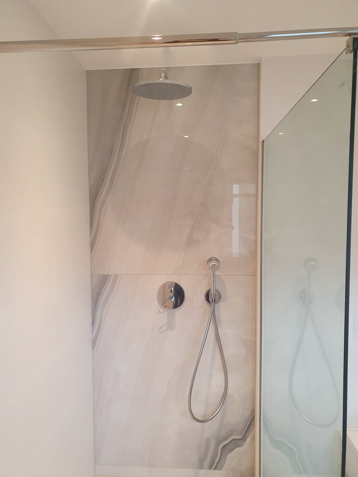 Wykończona kabina prysznicowa z deszczownicą i baterią, wyłożona płytkami imitującymi marmur. Szklane drzwi kabiny z widocznym odbiciem armatury.