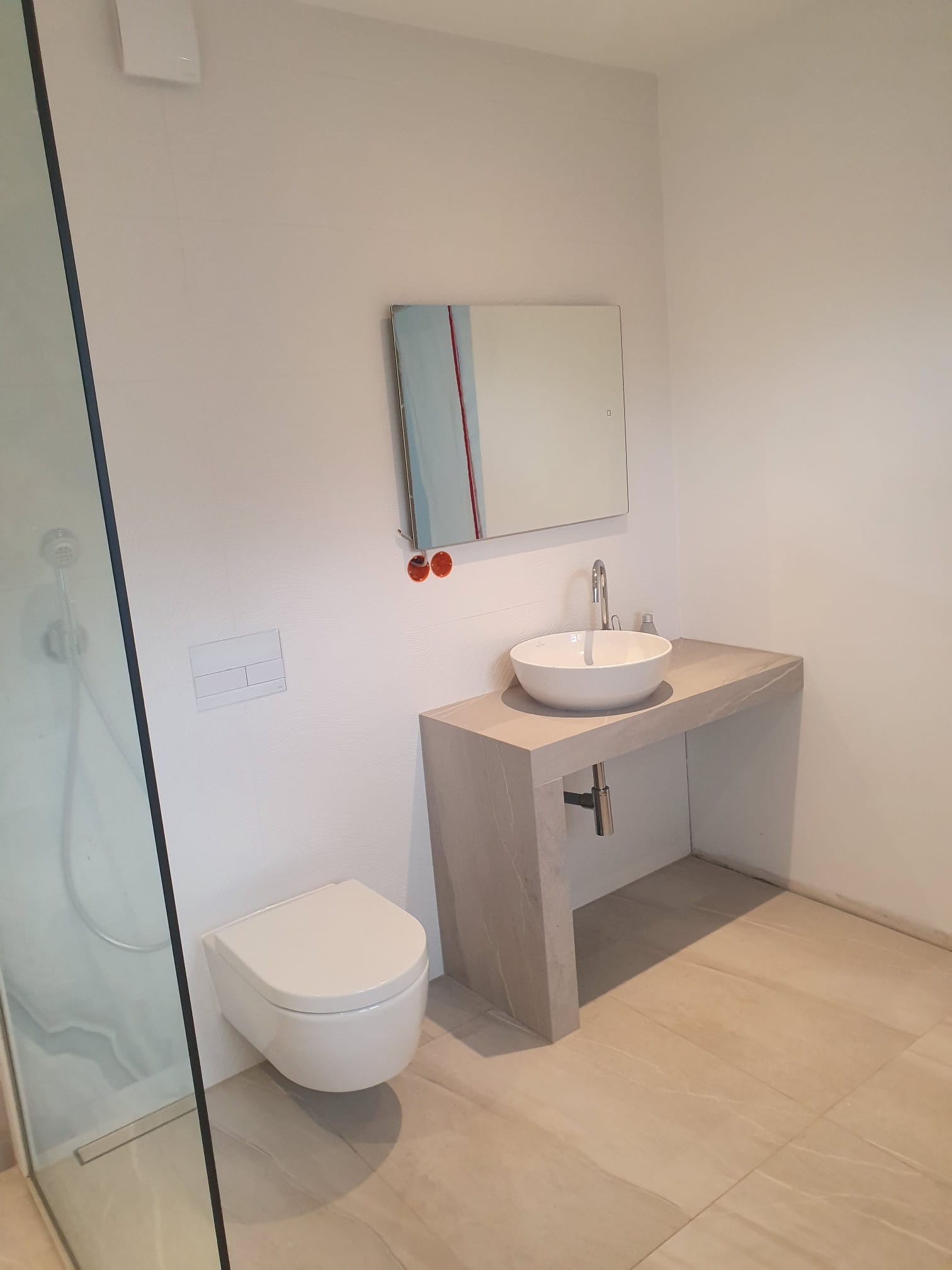 Nowoczesna łazienka z podwieszaną toaletą, umywalką nablatową na blacie z płytek imitujących marmur i minimalistycznym lustrem z akcentami kolorystycznymi.