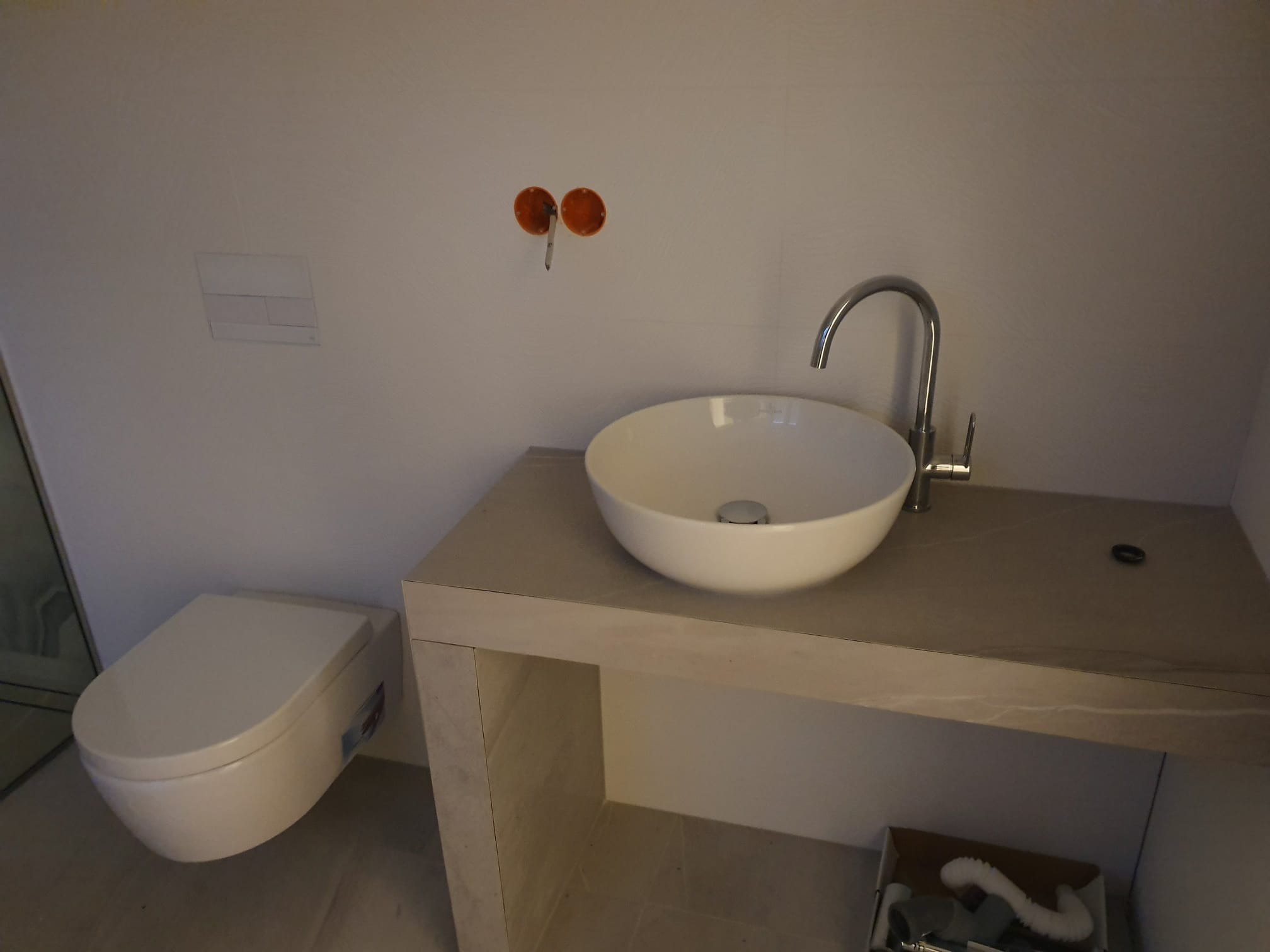 Nowoczesna łazienka w trakcie wykańczania: biała umywalka na blacie imitującym marmur, wisząca toaleta, widoczne instalacje. Minimalistyczny design.