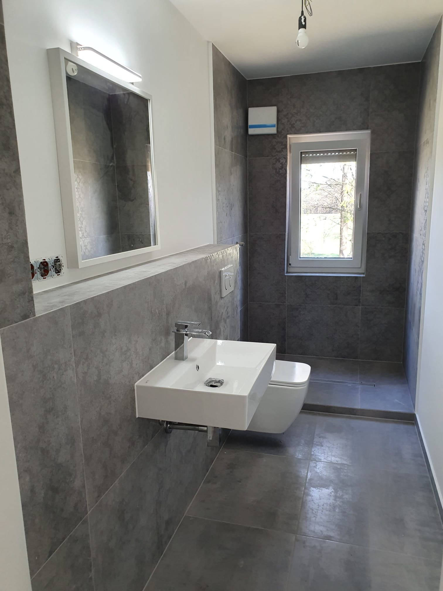 Nowoczesna łazienka w szarościach: minimalistyczna umywalka, lustro z oświetleniem, okno z widokiem na zieleń. Uwagę przykuwają płytki imitujące beton i surowy styl.