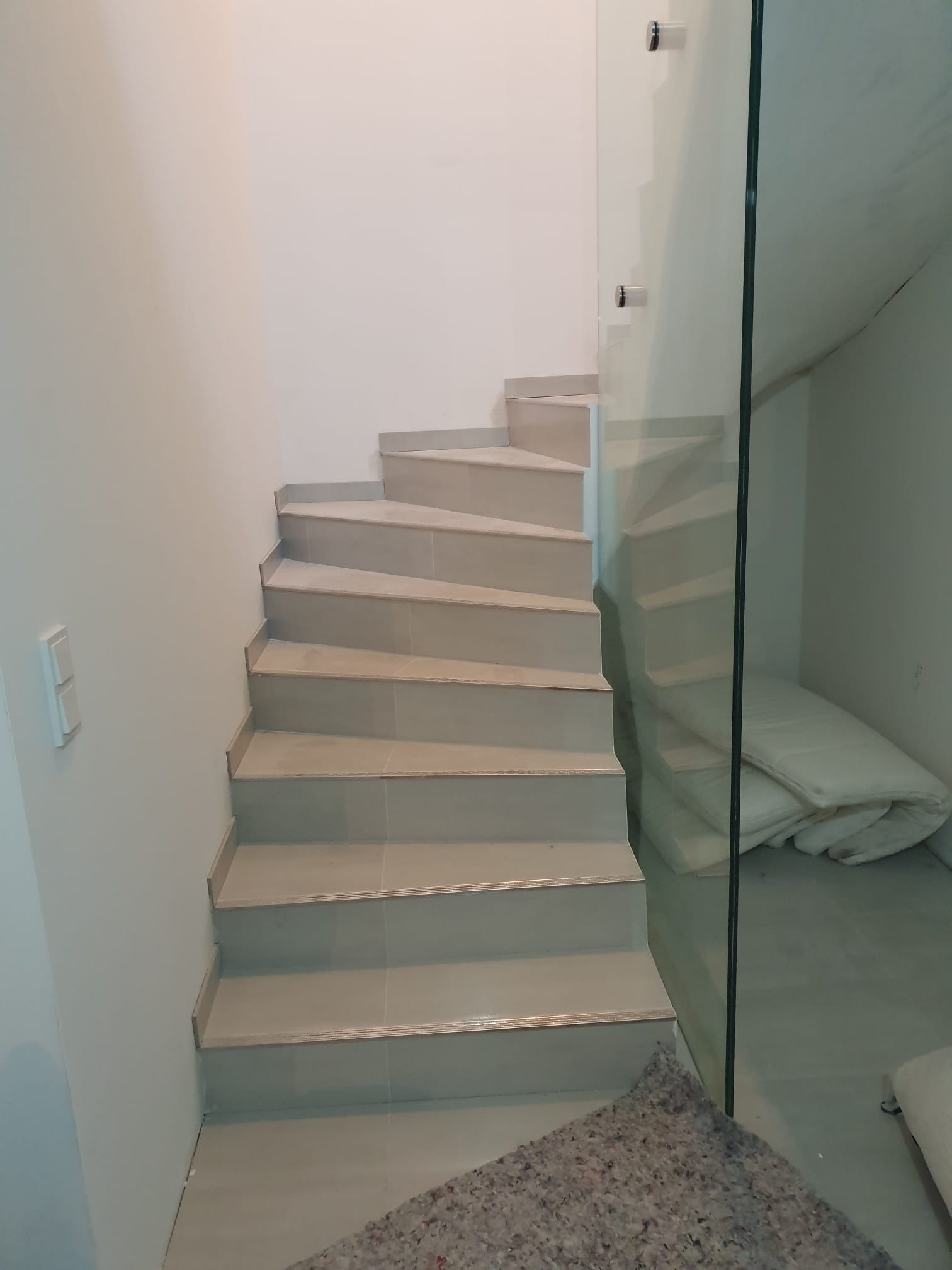 Spiralne schody wyłożone jasnymi płytkami, zabezpieczone szklaną balustradą. Widoczny fragment dywanu na dole schodów i materac za szybą.