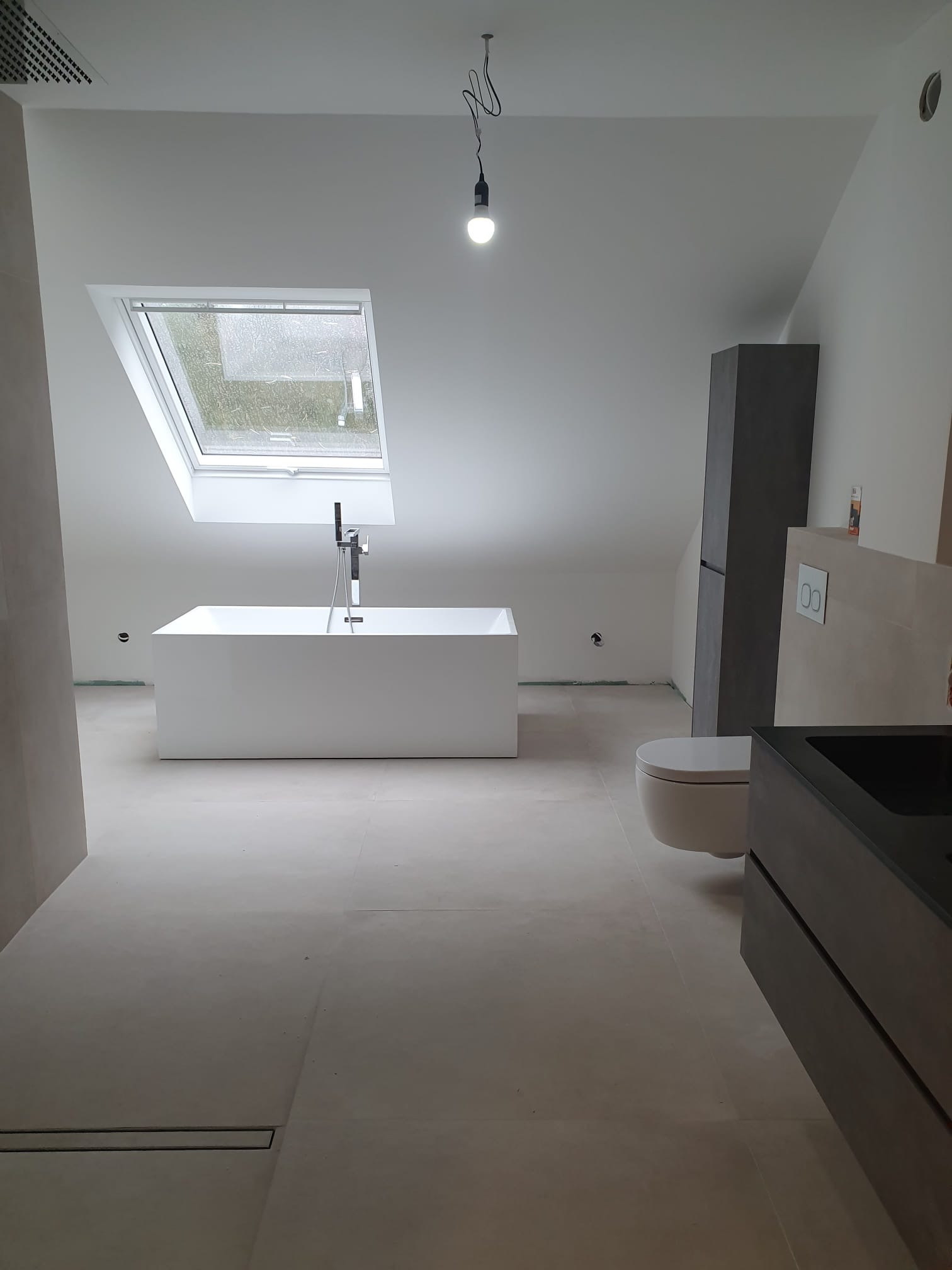 Nowoczesna łazienka na poddaszu z białą wanną wolnostojącą pod oknem dachowym, minimalistyczną toaletą i szafką z umywalką w odcieniach szarości.