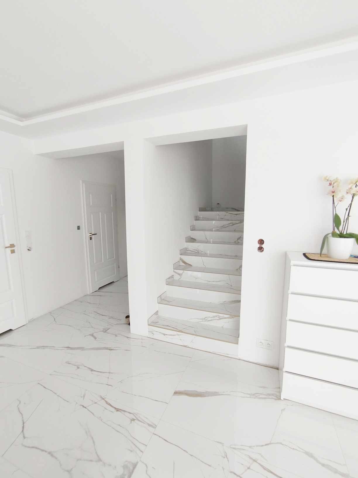 Białe wnętrze z marmurową podłogą i schodami. Minimalistyczny design, komoda z kwiatami, drzwi w tle. Jasne, przestronne pomieszczenie po remoncie.