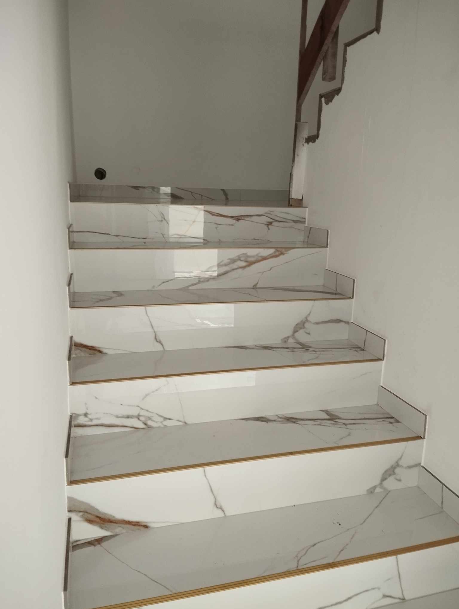 Wykończone schody z białych płytek imitujących marmur z delikatnymi złotymi akcentami, prowadzące w górę. Widoczne fragmenty drewnianej konstrukcji schodów.