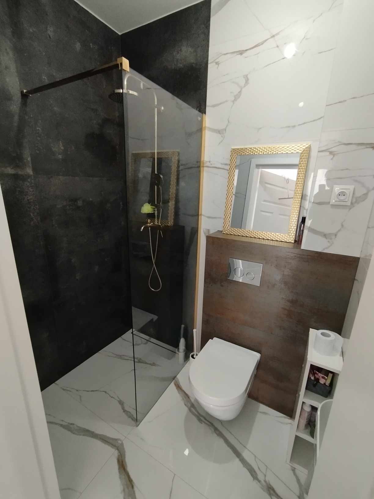 Nowoczesna łazienka z kabiną typu walk-in, czarną ścianą prysznicową i białymi płytkami imitującymi marmur. Złota rama lustra i akcenty kolorystyczne.