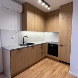 ArtHouse Raman Shkurko - Jasna kuchnia z drewnianymi frontami, czarnym zlewem i blatem imitującym beton. Nad blatem marmurowe wykończenie. Na suficie czarna lampa. Widoczny klimatyzator.