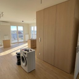 ArtHouse Raman Shkurko - Jasna szafa wnękowa z drewnianymi frontami w nowoczesnym mieszkaniu. Na podłodze pralka i narzędzia, sugerujące montaż. Widok na miasto z okna.