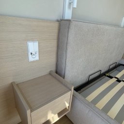 ArtHouse Raman Shkurko - Beżowe łóżko z szafką nocną z jasnego drewna, wbudowaną w wezgłowie. Nad szafką gniazdko i włącznik światła, a obok lampka nocna. Minimalistyczny design.