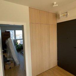 ArtHouse Raman Shkurko - Jasna, drewniana szafa wnękowa od podłogi do sufitu w przedpokoju, obok ciemnych drzwi wejściowych. Widoczne kartony i roślina w tle, sugerujące przeprowadzkę.