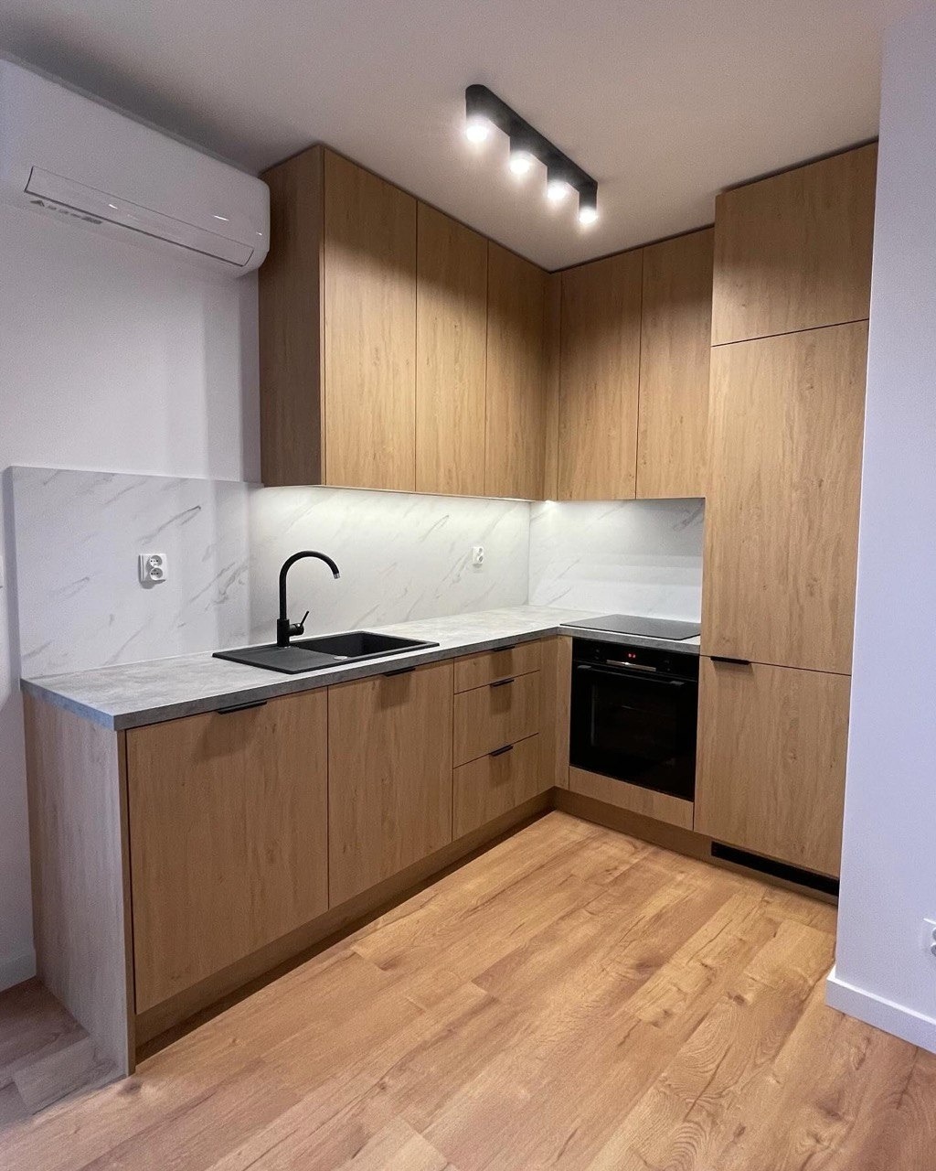 Jasna kuchnia z drewnianymi frontami, czarnym zlewem i blatem imitującym beton. Nad blatem marmurowe wykończenie. Na suficie czarna lampa. Widoczny klimatyzator.