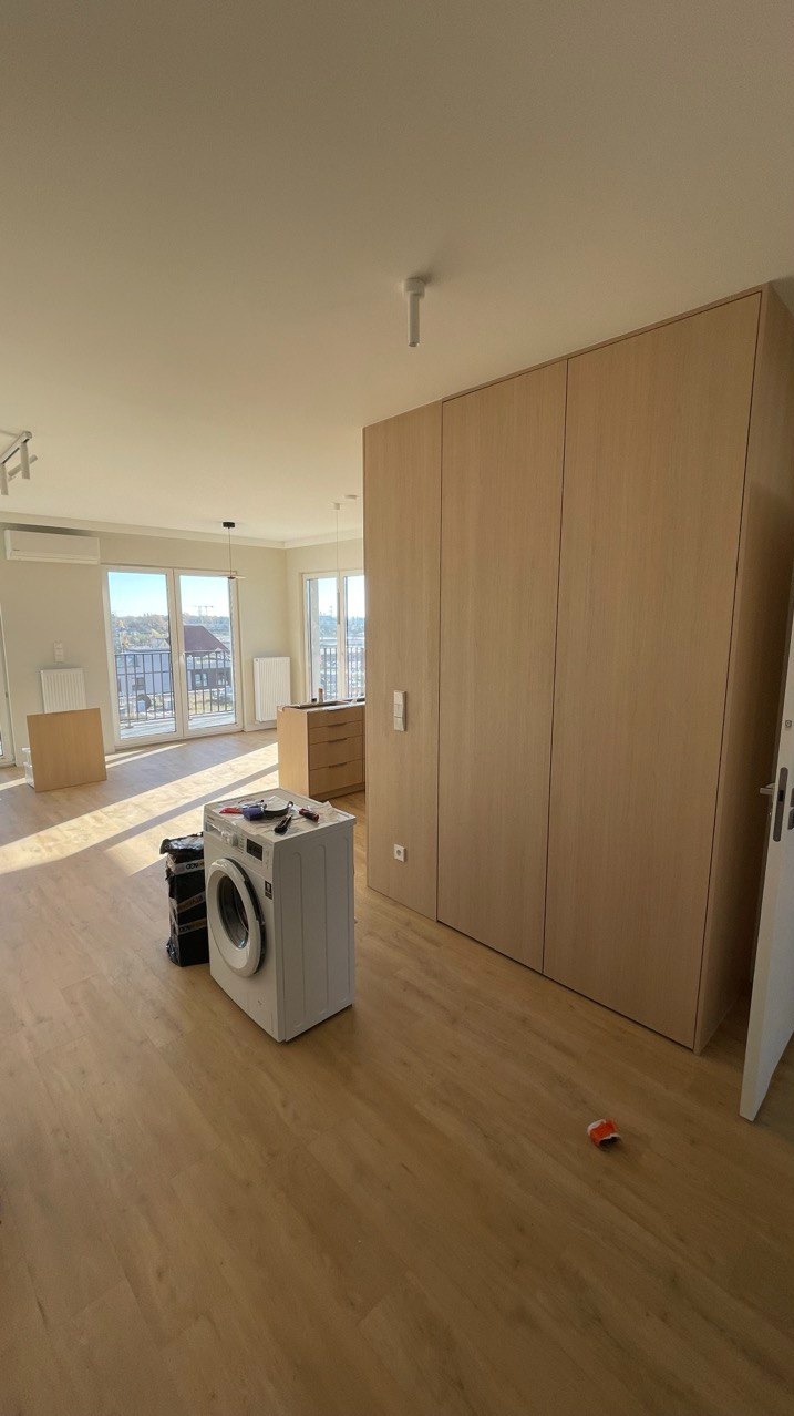 Jasna szafa wnękowa z drewnianymi frontami w nowoczesnym mieszkaniu. Na podłodze pralka i narzędzia, sugerujące montaż. Widok na miasto z okna.