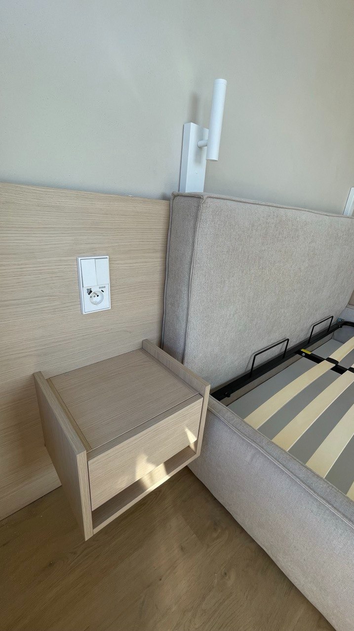 Beżowe łóżko z szafką nocną z jasnego drewna, wbudowaną w wezgłowie. Nad szafką gniazdko i włącznik światła, a obok lampka nocna. Minimalistyczny design.