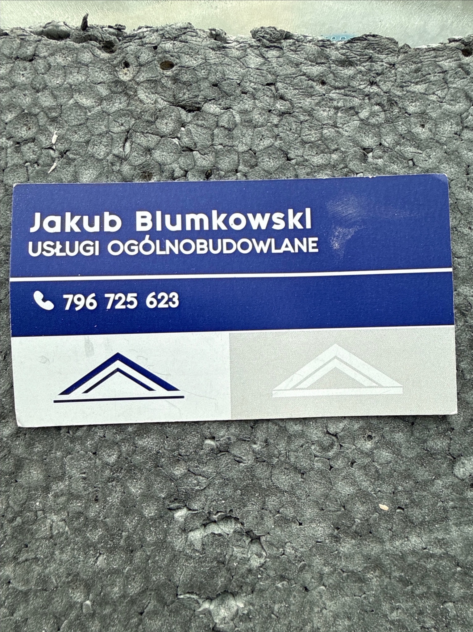 Wizytówka Jakuba Blumkowskiego, usługi ogólnobudowlane, numer telefonu na tle szarej, chropowatej powierzchni, prawdopodobnie styropianu.