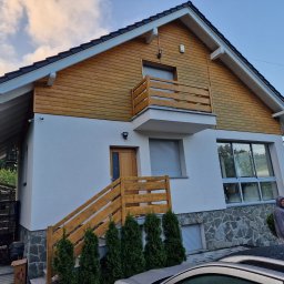 Usługi ogólnobudowlane PROHOUSE - Dwukondygnacyjny dom z drewnianą elewacją na poddaszu i białymi ścianami. Widoczne drewniane schody wejściowe i balkon. W tle zalesione wzgórza.