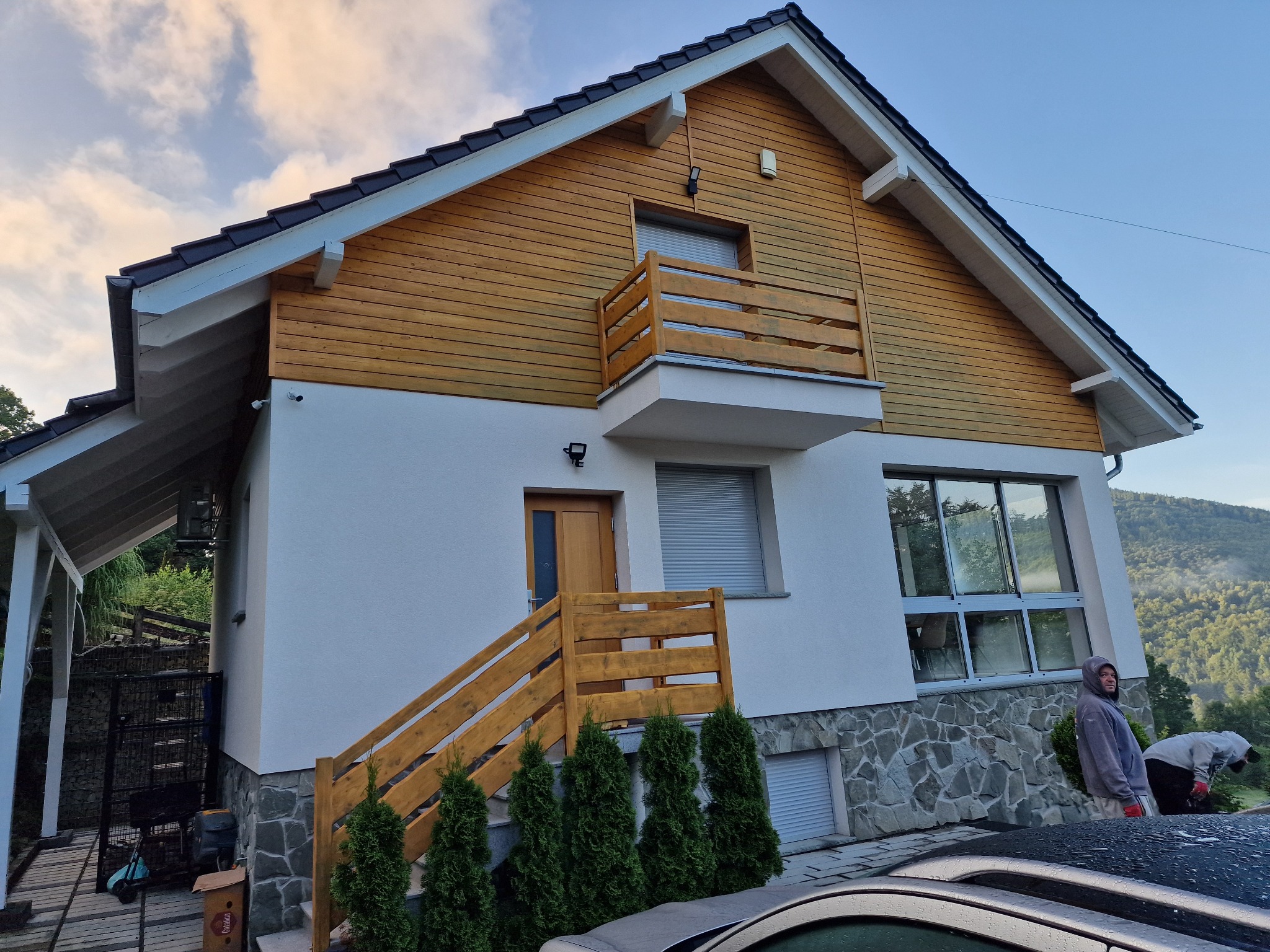 Dwukondygnacyjny dom z drewnianą elewacją na poddaszu i białymi ścianami. Widoczne drewniane schody wejściowe i balkon. W tle zalesione wzgórza.