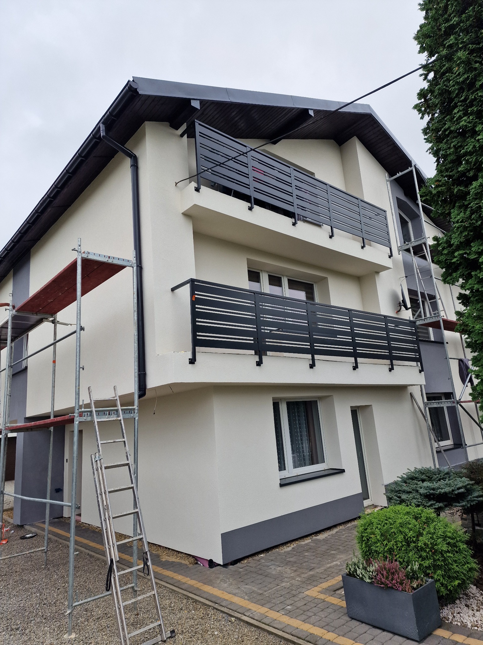 Dwukondygnacyjny dom z balkonami po renowacji elewacji w Brennej. Widoczne rusztowania i drabina, podkreślające etap prac wykończeniowych. Nowoczesne, czarne balustrady.