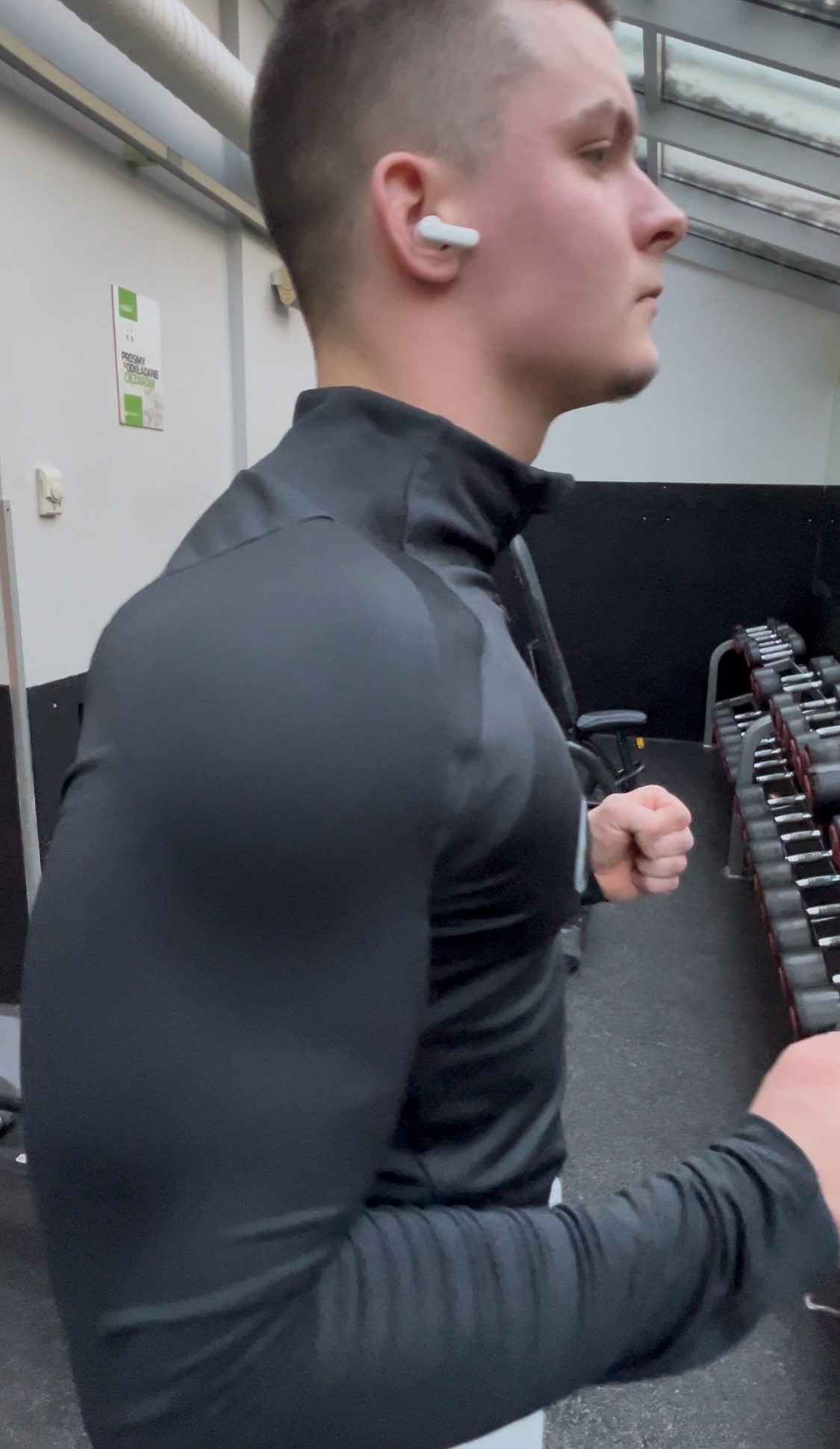 Mężczyzna w czarnej bluzie, z słuchawką w uchu, ćwiczy na siłowni, widoczne hantle w tle. Prezentacja sylwetki podczas treningu.