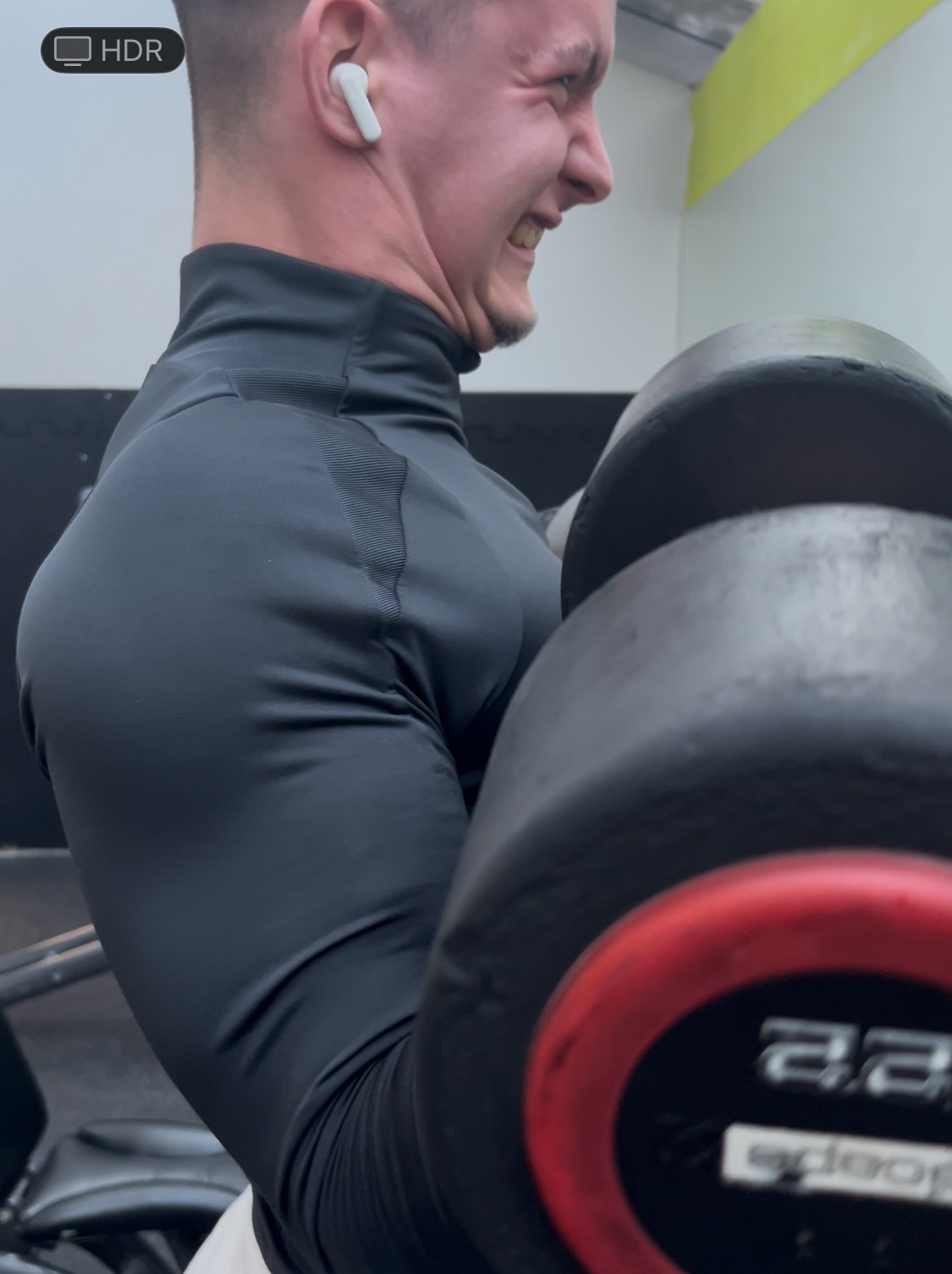 Mężczyzna w czarnej koszulce z długim rękawem ćwiczy biceps z ciężarem 22 kg. Widoczny wysiłek na twarzy, słuchawka w uchu. Ujęcie z bliska, z boku.