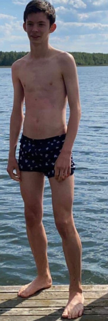 Młody mężczyzna w kąpielówkach stoi na drewnianym pomoście nad jeziorem w słoneczny dzień. Widoczna sylwetka, w tle woda i zieleń.