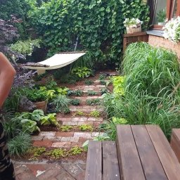 Janex - Ogród z ceglaną ścieżką, hamakiem i bujną roślinnością. Widoczne drewniane schody i fragment osoby. Kompozycja zróżnicowana, z elementami dekoracyjnymi.
