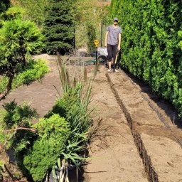 Janex - Wykopane rowy w ziemi pod system nawadniania ogrodu, widoczna osoba z łopatą i narzędzia ogrodnicze. Tuje w tle. Słoneczny dzień.