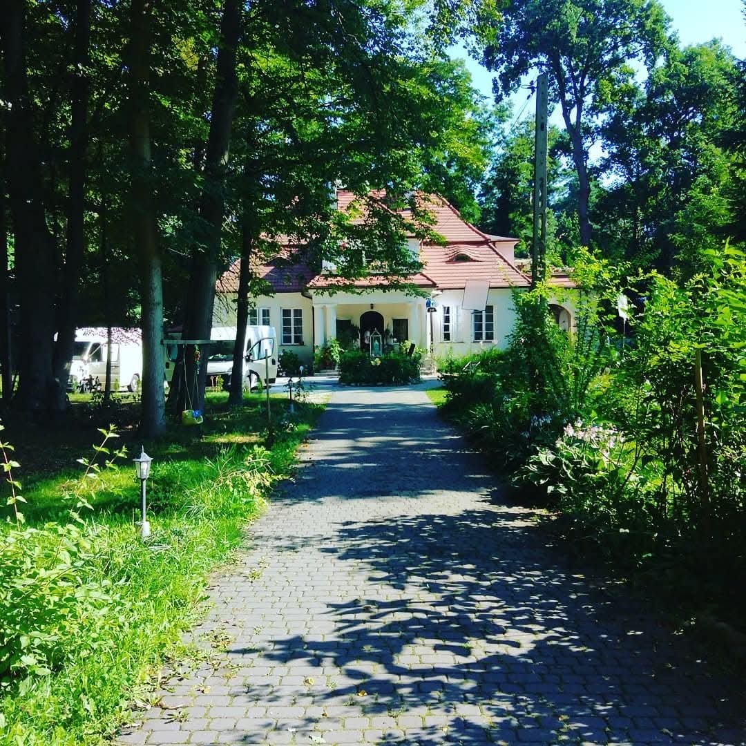 Brukowana ścieżka prowadzi do białego domu z czerwonym dachem, otoczonego zielenią drzew i krzewów w słoneczny dzień. Widoczne lampy ogrodowe i zaparkowane pojazdy.