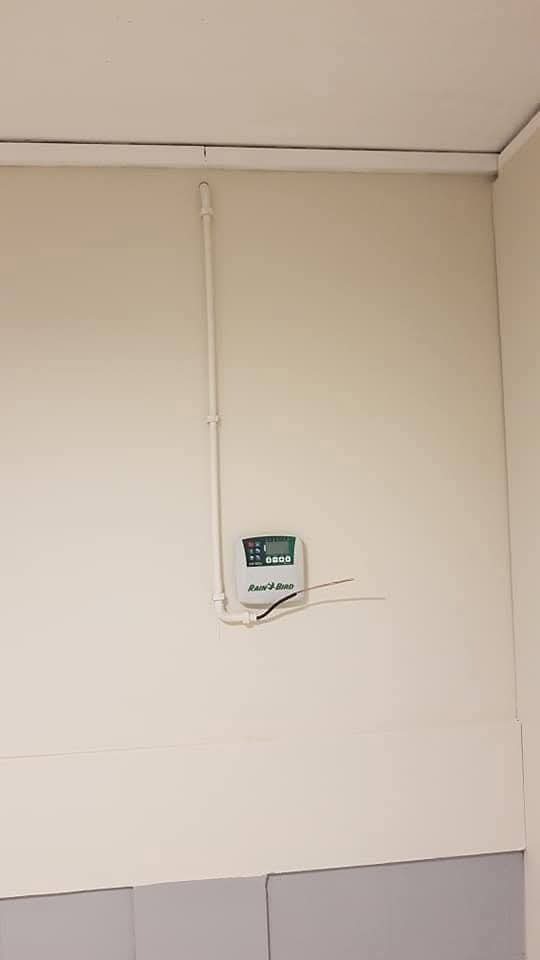 Zamontowany na ścianie sterownik systemu nawadniania Rain Bird z doprowadzonymi rurami. Widoczne okablowanie i fragment ściany z szarą lamperią.