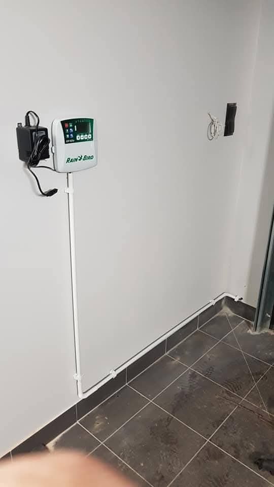 Zamontowany sterownik nawadniania Rain Bird na białej ścianie, z widocznymi przewodami i zasilaczem. Szare płytki podłogowe i fragment drzwi w tle.
